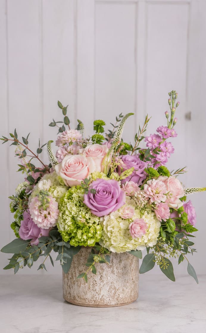 Perfectly Petal Perfection  - A lush, garden-style bouquet in soft blush pinks, lavender purples, crisp white and fresh greens-this flower arrangement is a romantic choice for a wedding bouquet alternative, anniversary roses gift, or a springtime surprise. The rounded composition feels intentionally abundant and gentle, the kind of arrangement that makes the person you're thinking of pause and smile at its soft colors and layered textures.  Large roses anchor the design while voluminous hydrangea clusters and delicate stock and other small blooms build a plush, textural bouquet. Airy branching greenery adds height and movement without stiffness, creating a softly tiered profile that reads beautiful from every angle. The flowers come nestled in a textured ceramic vase that complements the palette and makes the arrangement ready to display on a dining table, mantel, or bedside table.  Perfect for celebrating an anniversary, saying "I'm thinking of you," or dressing a spring event, this arrangement delivers a romantic, fresh impression your recipient will notice. Vase included; arrange in a cool spot away from direct heat and change the water every 2-3 days to keep the roses and hydrangeas looking their best.