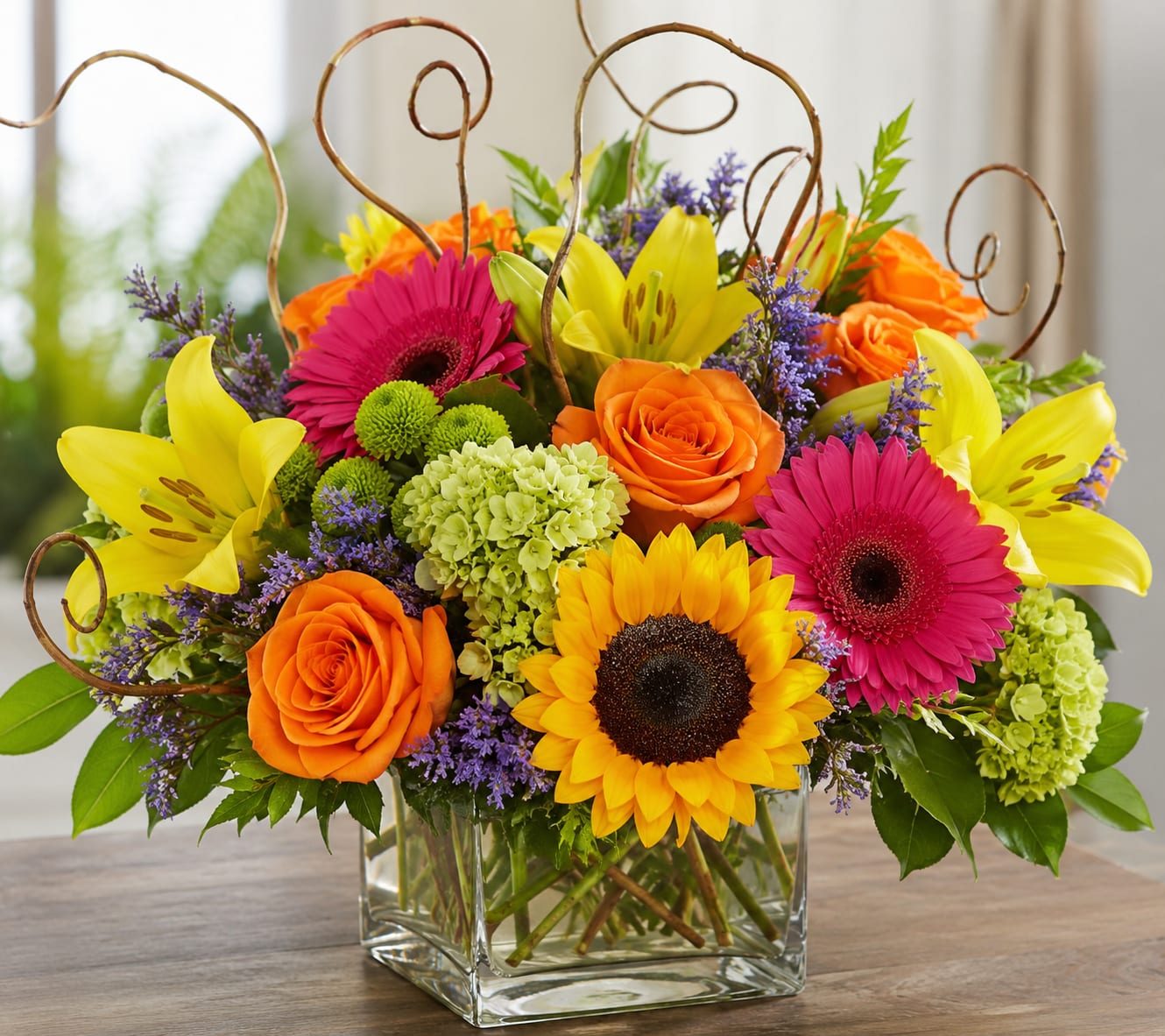Summer Time Sunshine  - A bold sunflower crown pulls the eye in-this Summer Time Sunshine flower arrangement is a cheerful bouquet perfect for a birthday bouquet, summer celebration, or "just because" surprise. Bright orange roses and hot pink gerbera daisies mingle with yellow lilies to create a sunlit, uplifting look that says celebration; the composition reads as energetic and joyful for anyone you want to brighten.  The clear square glass vase holds a lively mix of blooms: a dominant central sunflower, orange roses, hot pink gerbera daisies, yellow lilies, small green hydrangea clusters, and purple filler flowers for contrast. Decorative curled twigs rise above the arrangement to add height and a playful, airy silhouette. Colors include pink, orange, yellow, and purple for a warm-weather palette that pops in any room.  This arrangement is ideal for sending to someone who should feel noticed and celebrated-the kind of bouquet that makes the recipient smile the moment it arrives. Vase-arranged for easy display, simply trim stems and refresh water every few days to extend the life of the flowers.