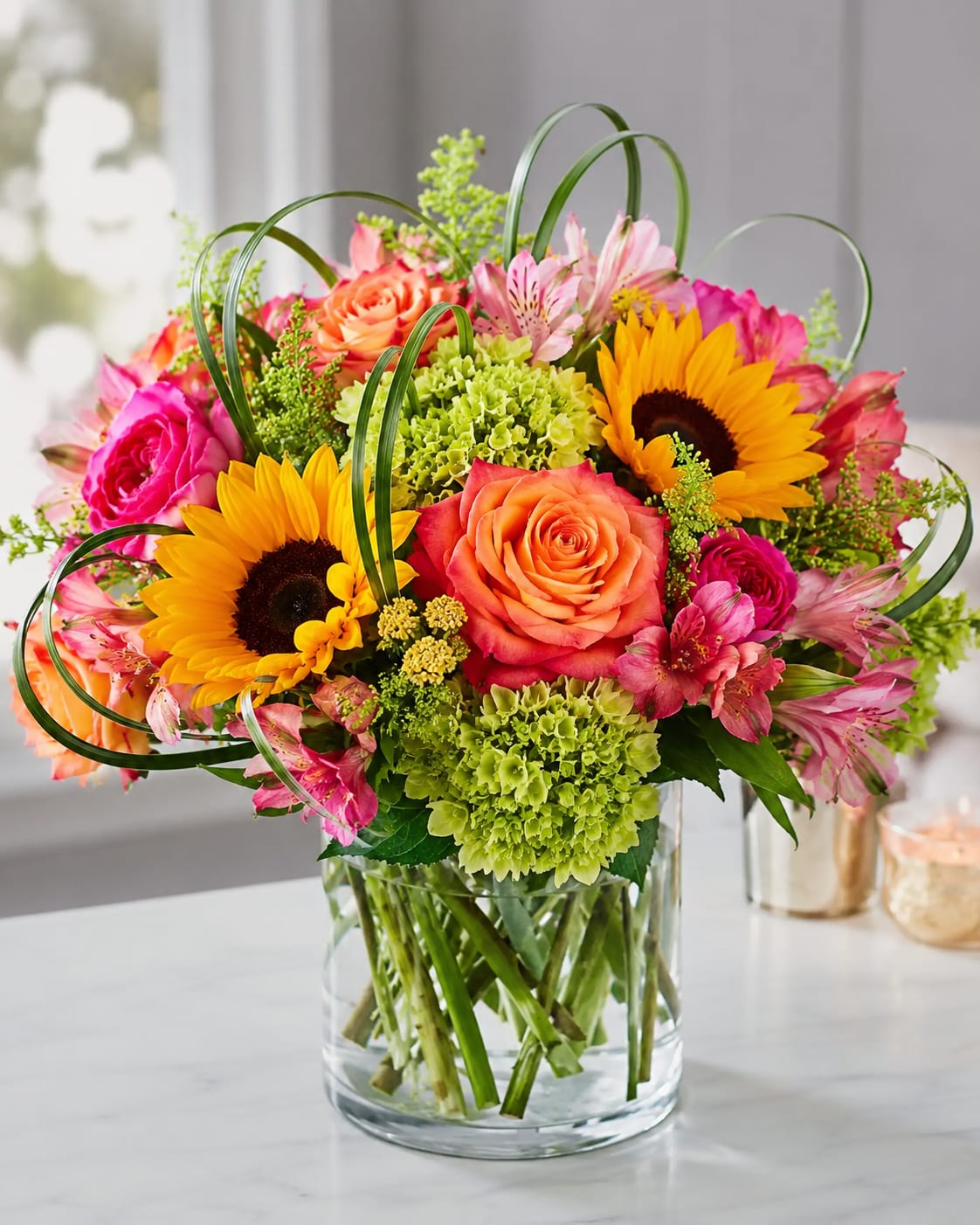 Dancing in the Sun - Bright yellow sunflowers and coral-pink roses give this birthday bouquet a sunny, celebratory feel that instantly lifts the room-perfect for birthdays, congratulations, or a cheerful thank-you gift. The mix of pink alstroemeria and green hydrangea clusters keeps the palette fresh and garden-like, so the recipient will feel genuinely seen and brightened by the lively color play.  Arranged in a clear glass vase, Dancing in the Sun pairs saturated yellow, orange, pink, and green blooms with curving bear grass for a loose, airy structure that reads contemporary and playful. The combination of bold sunflower faces, soft rose petals, delicate alstroemeria blooms, and textured hydrangea gives the bouquet visual depth and a tactile contrast that's as pretty up close as it is from across the room.  This everyday celebratory arrangement works wonderfully for friends, coworkers, or anyone who could use a joyful pick-me-up; it delivers an upbeat, optimistic message without being over the top. Vase-ready and easy to display, it arrives arranged and ready to enjoy-simple care (fresh water and a cool spot) will keep the blooms looking their best.