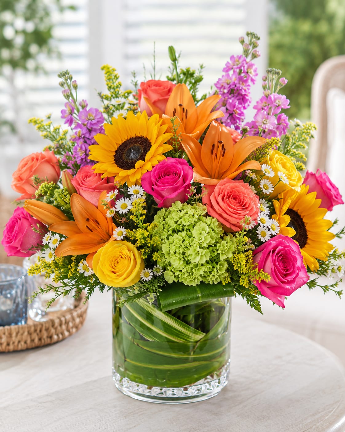 Summer Grand by Petal Perfection - A burst of summer sunshine and garden charm greets the recipient with this mixed-color flower arrangement for celebrations and uplifting moments. For a birthday bouquet, get-well surprise, or just-because gift, the Summer Grand by Petal Perfection delivers bold focal blooms and playful texture that will make the person you're thinking of feel truly celebrated.  Sunflowers and orange lilies form a bright, confident center while hot pink and coral roses soften the composition with romantic warmth. Yellow hydrangea tones and small white daisy-like fillers add lively contrast and a meadow-like texture, all arranged in a clear glass vase that lets the colors sing. The dense, rounded silhouette reads generous and joyful from every angle.  Perfect for sending congratulations, cheering someone up, or marking a sunny anniversary, this arrangement arrives ready to display. Expect a substantial, garden-style bouquet that brings energetic color to tabletops, desks, or entryways; refresh the water every few days and trim stems to extend its cheerful presence.