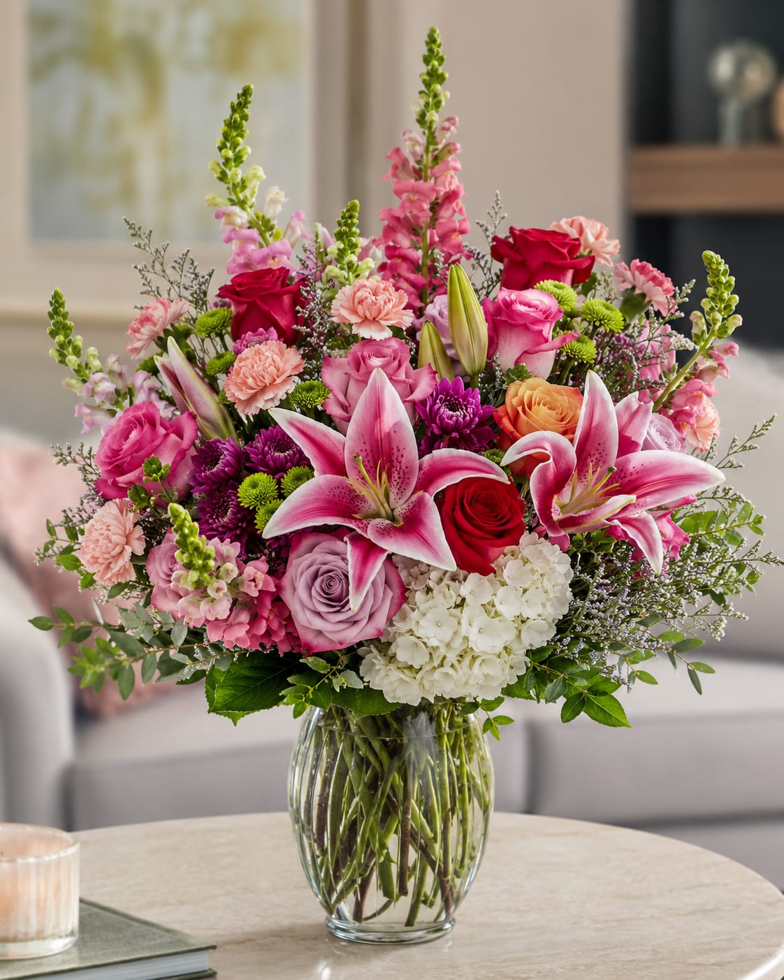 Petal Perfection's You're Precious To Me - Bright pink stargazer lilies take center stage in this large, lush flower arrangement-an abundant mixed bouquet perfect for a birthday bouquet, anniversary flowers, or Mother's Day surprise. The palette of pink, white, purple, and red creates a bright, romantic impression that reads celebratory but personal; the person you're thinking of will feel truly cherished when greeted by these soaring lilies and soft rose blooms.  This upright garden-style design pairs red, pink, lavender and peach roses with white hydrangea, clusters of carnations and chrysanthemums, and tall snapdragon spikes for height and movement. Arranged in a clear glass vase, stems are gathered tightly below the blooms for a full, layered look that showcases texture and color from every angle. The mix of large lily blooms, rounded hydrangea heads, and vertical snapdragons creates both visual drama and a plush, touchable feel.  Ideal for marking special moments-Mother's Day, anniversaries, milestone birthdays-or simply to say "you're precious to me," this arrangement reads as thoughtful and generous without being fussy. Size and presence make it a focal gift for a dining table or entryway; to keep it looking fresh, change the water every 2-3 days and trim stems slightly when refreshing the vase.
