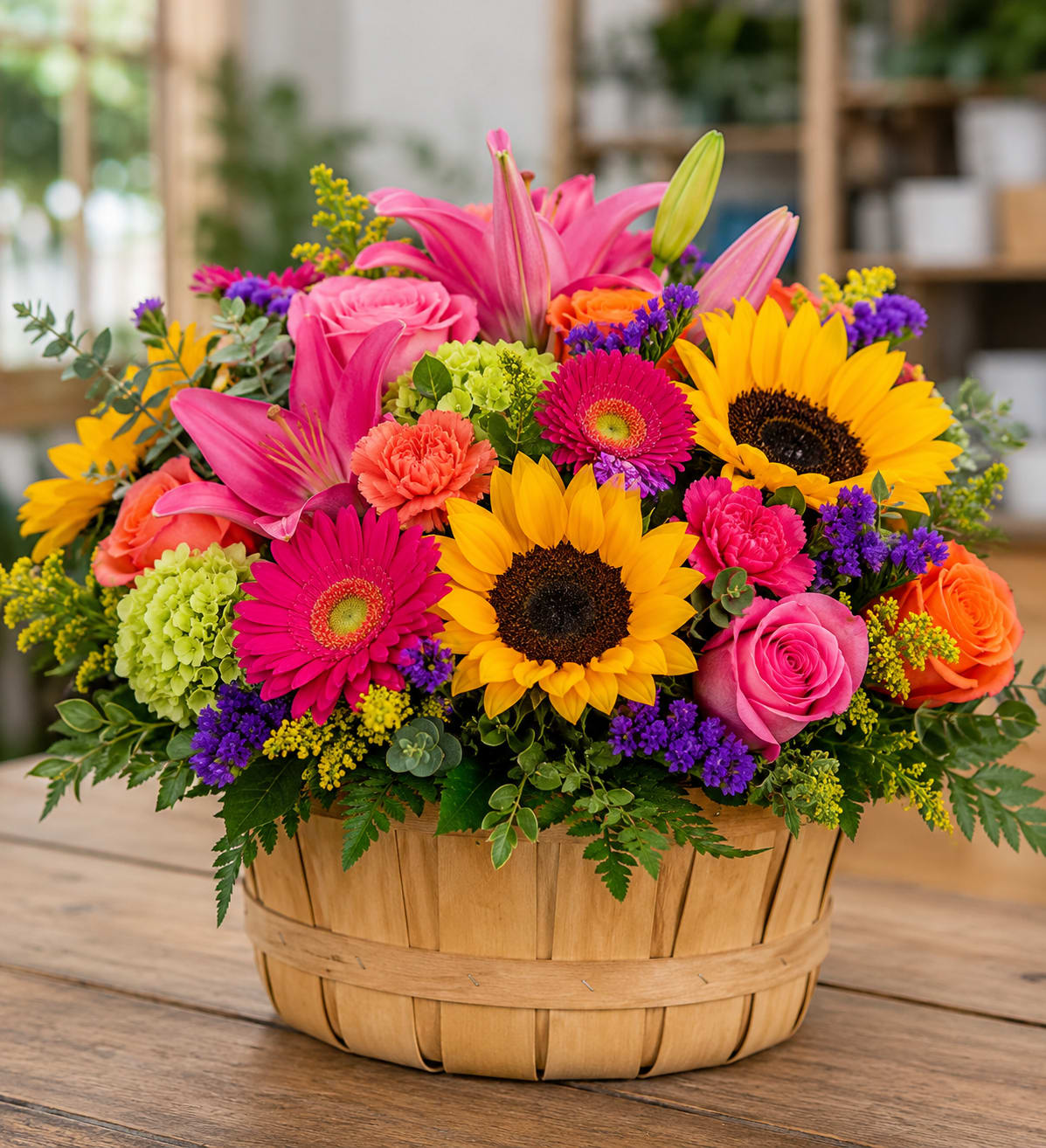 Bonnie Basket for Ruskin  - Bright yellow sunflowers and hot pink gerbera daisies make a bold, cheerful statement in this flower arrangement-perfect as a birthday bouquet, congratulations gift, or a bright everyday surprise. The woven wooden basket gives the composition a warm, rustic feel while the mix of pink, yellow, purple, and orange blooms creates an immediate sense of celebration that will make the person you're thinking of feel truly seen and uplifted.  This large, colorful mixed bouquet features prominent sunflowers, lilies, pink and orange roses, gerbera daisies, carnations, and green hydrangea-like blooms arranged densely for a lush, layered look. Small purple filler flowers and assorted greenery add contrast and texture around the edges, so the arrangement reads lively from every angle. The wooden basket provides sturdy presentation and easy placement on a table or countertop.  Ideal for sending congratulations, brightening a birthday morning, or saying "thinking of you" with extra cheer, this basket arrives ready to display-no vase required. To keep the flowers looking their best, place the arrangement in a cool spot away from direct sunlight and refresh the water if the basket's liner allows; sizing is generous, designed to create a joyful focal point in any room.