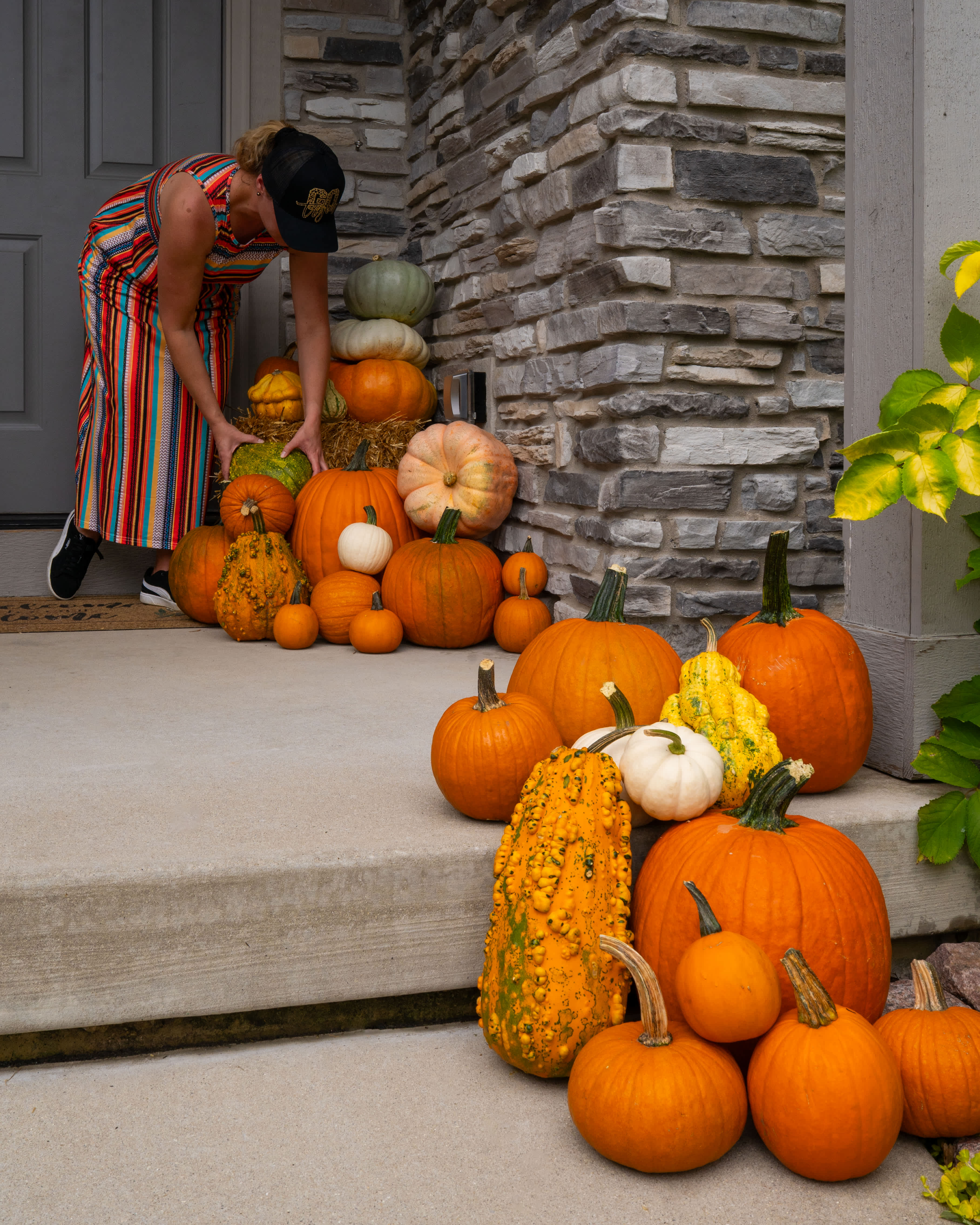 A woman arranges a stack of pumpkins and gourds on a front porch.