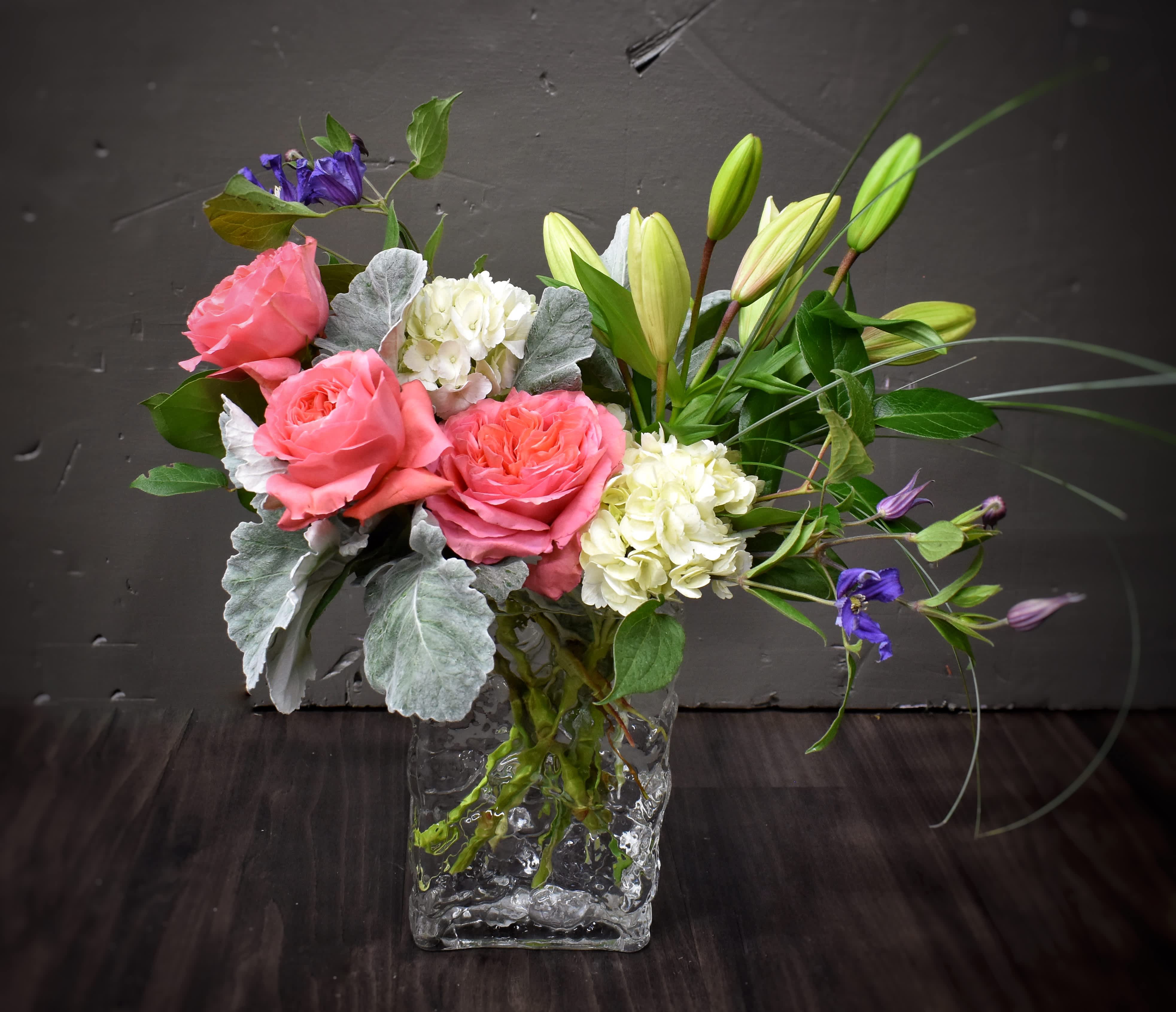 Arrangement of pink roses, white hydrangeas, and lilies in a clear glass cube vase with purple accents