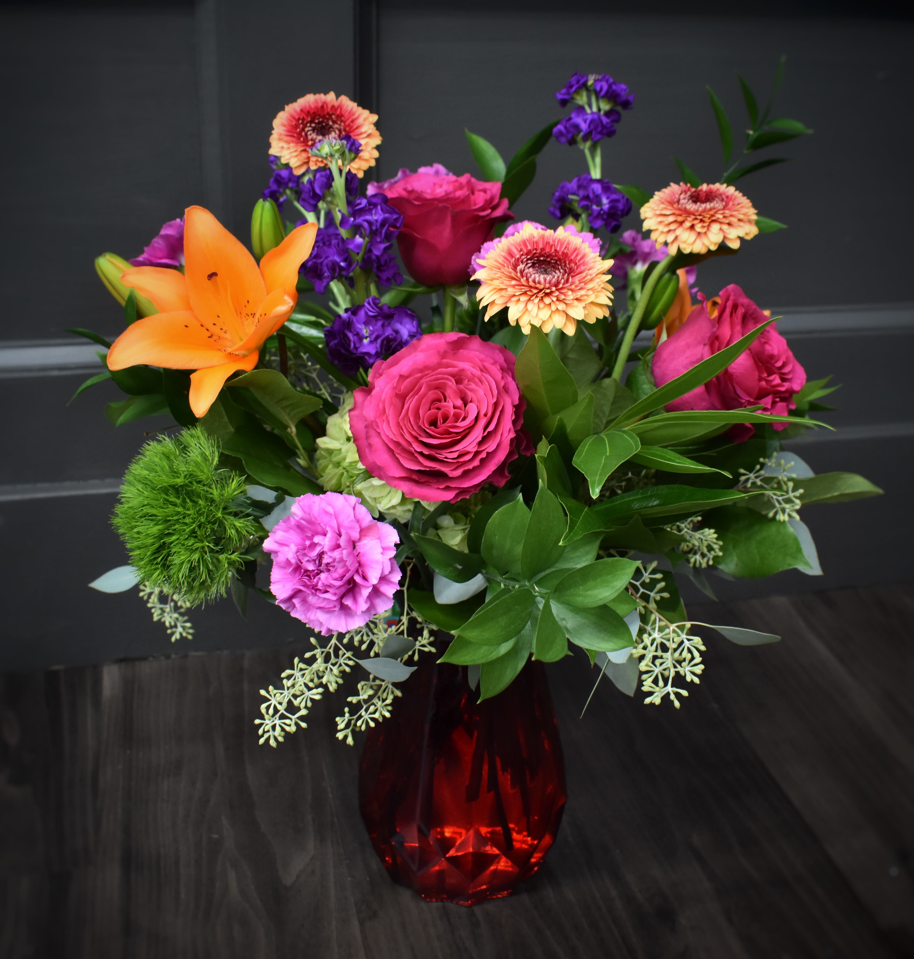 Mixed bouquet of roses, lilies, and gerbera daisies in a red glass vase