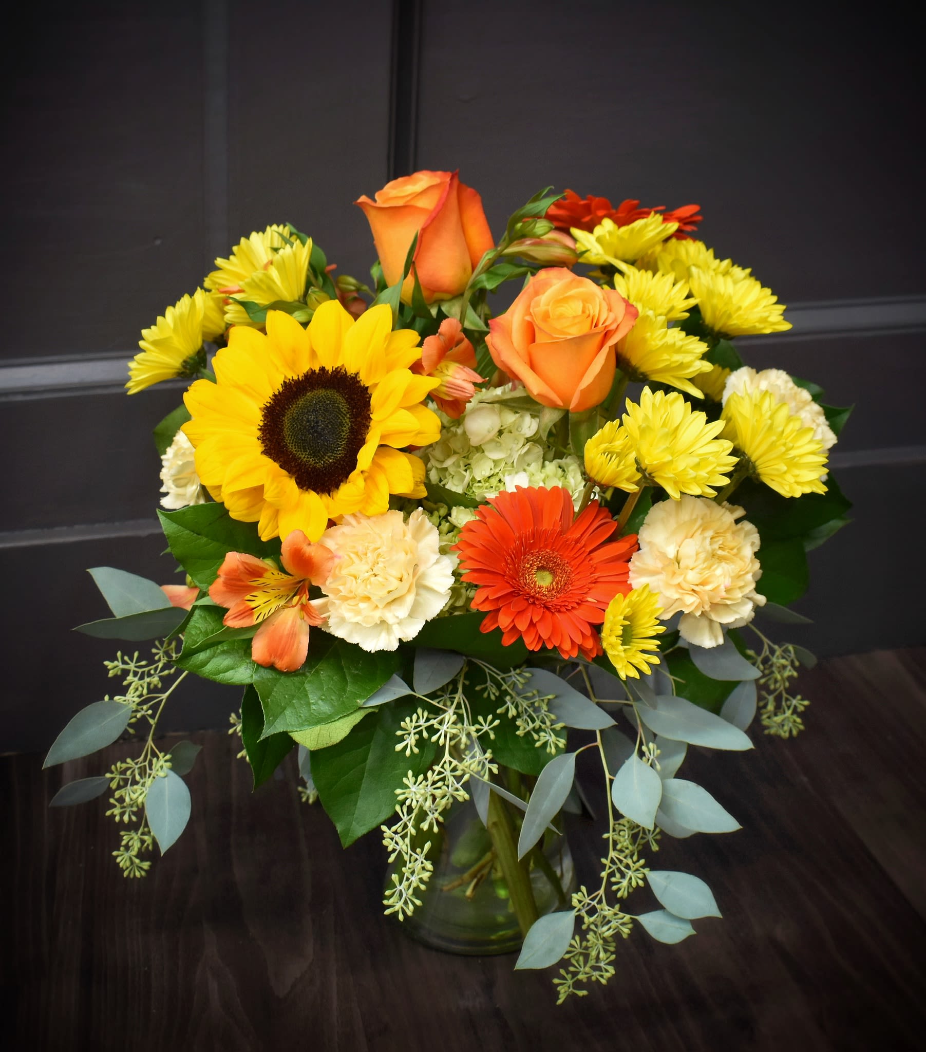 Mixed bouquet with sunflowers, orange roses, gerbera daisies, and yellow mums in a glass vase