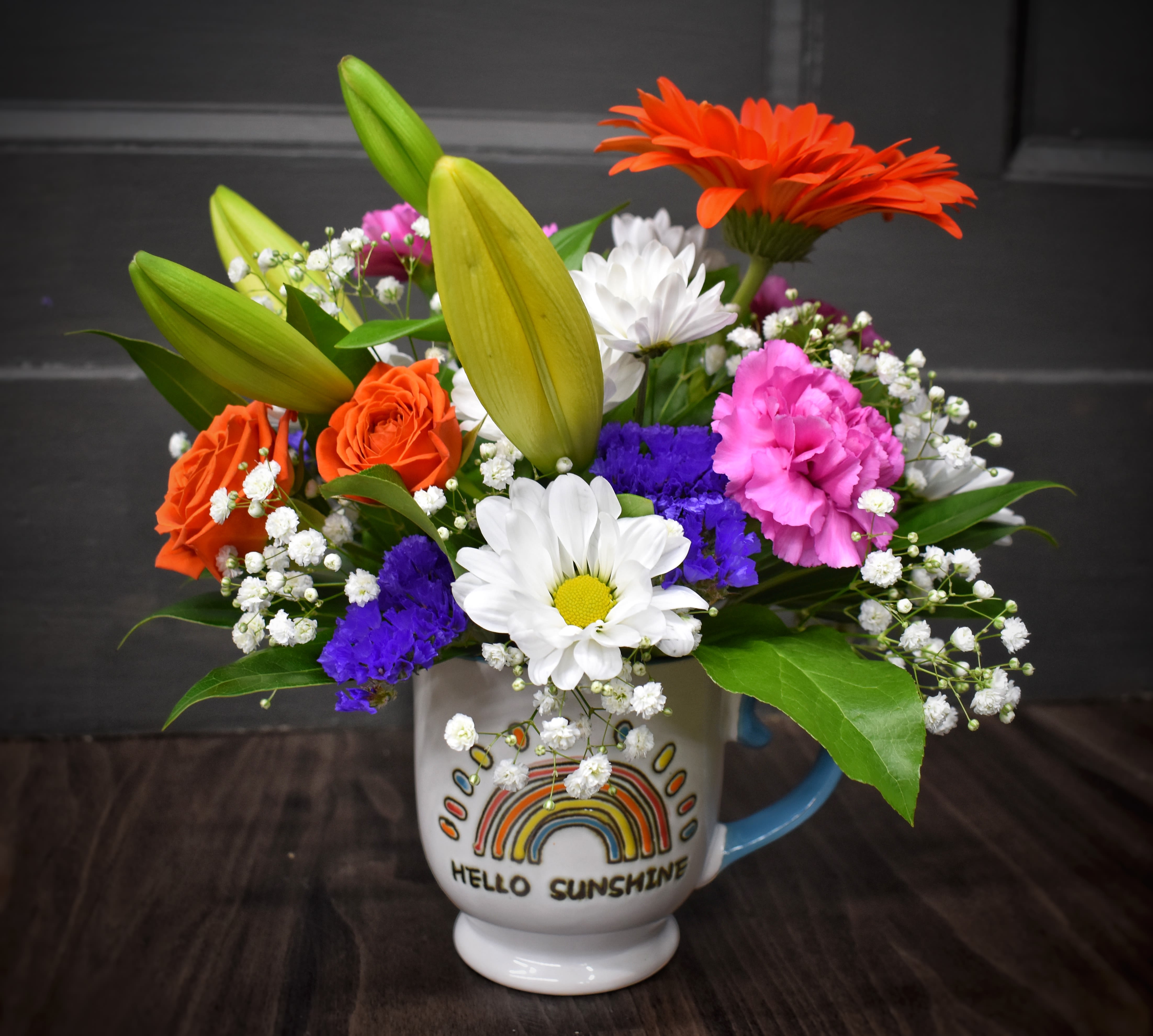 Hello Sunshine Mug Bouquet - Bright orange roses and gerbera daisies pop against soft pink carnations, white daisies, and sprays of purple filler in this Hello Sunshine mug bouquet - a cheerful flower arrangement perfect for a birthday, a thank-you, or a spontaneous pick-me-up. Unopened lily buds reach above the mix to add height and a hint of fragrance, so the person you're sending it to will feel noticed and uplifted the moment the mug arrives.  The palette of orange, pink, white, blue accents, and mixed filler (including tiny white baby's breath) creates a layered, playful look. The bouquet is delivered in the printed ceramic mug, making it both vase and keepsake - easy to water and enjoy again and again.
