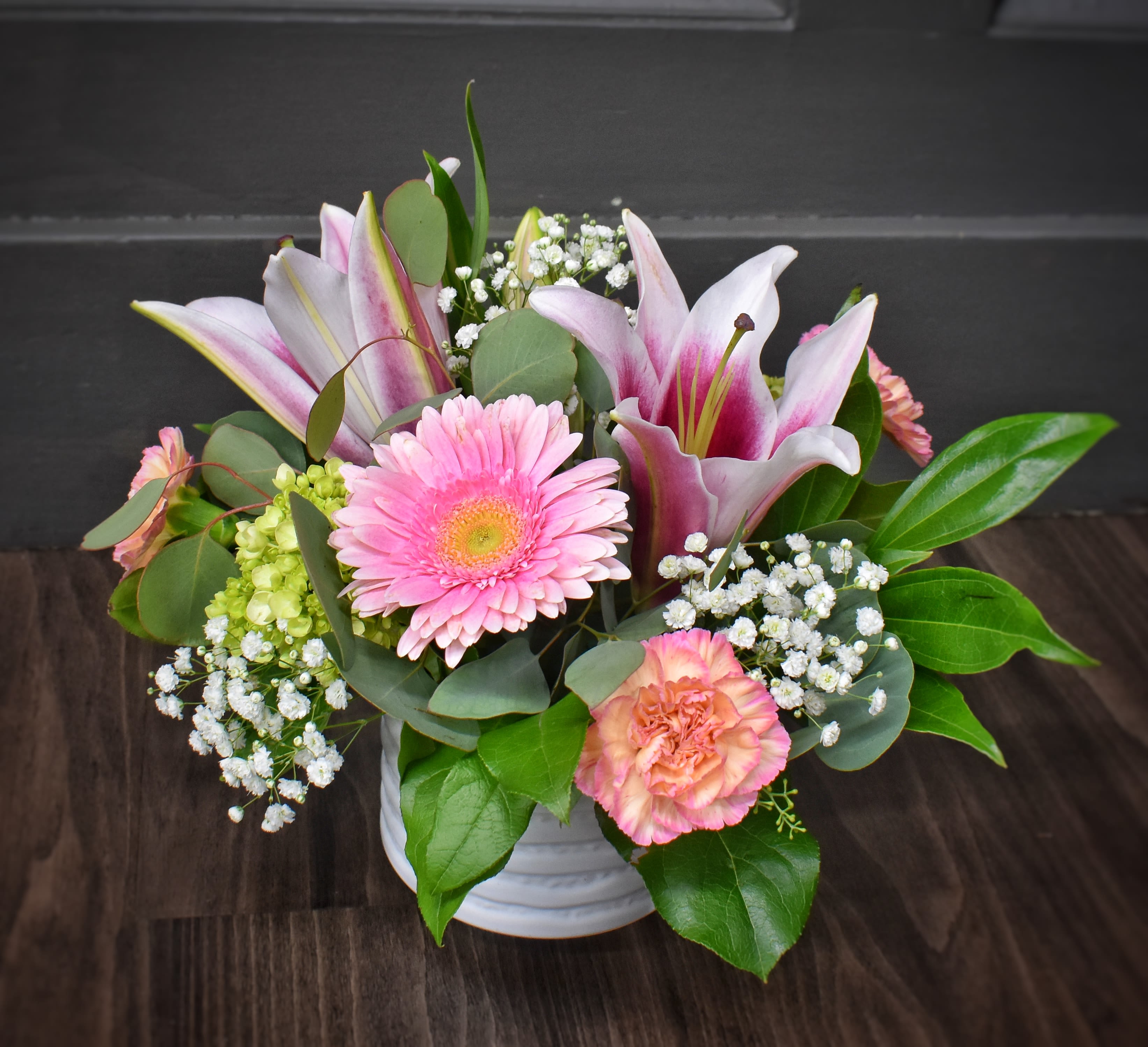 Pink Pixie - A bright cluster of pink lilies and  large pink gerbera daisies give this flower arrangement an immediate, joyful focal point-perfect as a birthday bouquet, a thank-you gift, or a cheerful everyday surprise. The Pink Pixie arrangement reads feminine and fresh, sending a message of warmth and celebration that will make the person you're thinking of feel seen and smiled at.  Soft peach-pink carnations, pale pink-and-white lilies, and bright pink roses mingle with small white filler blooms, rounded eucalyptus-like foliage, and a touch of green hydrangea to add texture and contrast. The compact, rounded design sits in a low white ceramic vase for a polished tabletop presence; its glossy leaves and layered petals create a garden-style look that feels both elegant and casual.  Ideal for desks, kitchen tables, or as a centerpiece for small gatherings, Pink Pixie is sized to fit most surfaces without overwhelming them. Care is simple-keep the vase filled with fresh water and trim stems slightly every few days to prolong bloom life-so the recipient can enjoy its cheerful colors for as long as possible.