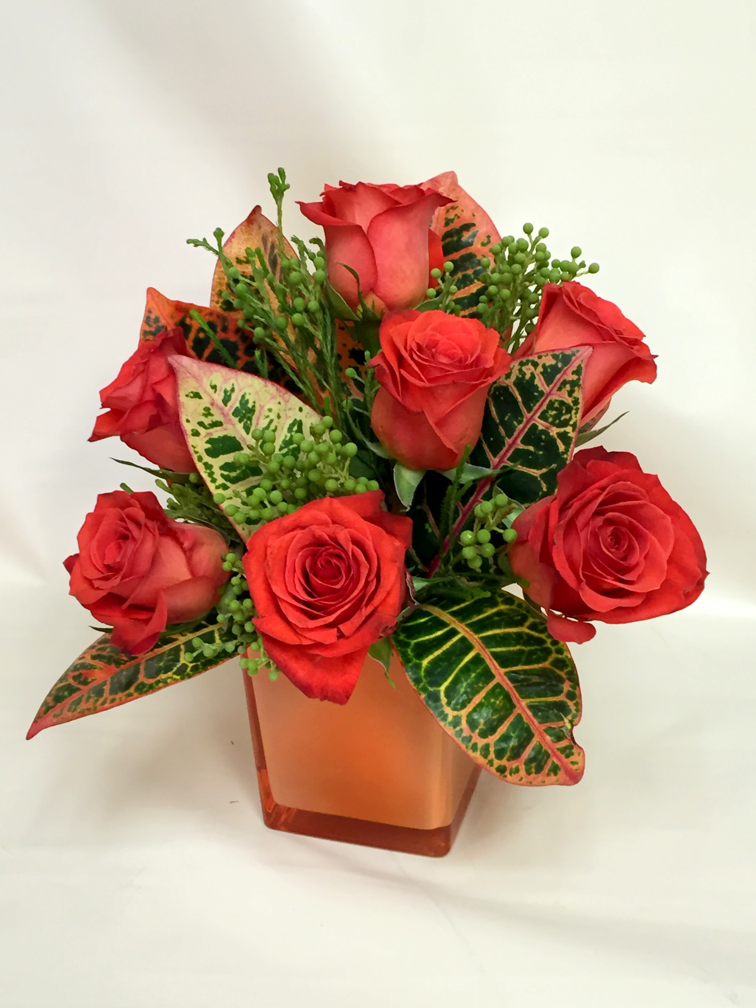 Red roses arranged in an orange square vase with variegated leaves