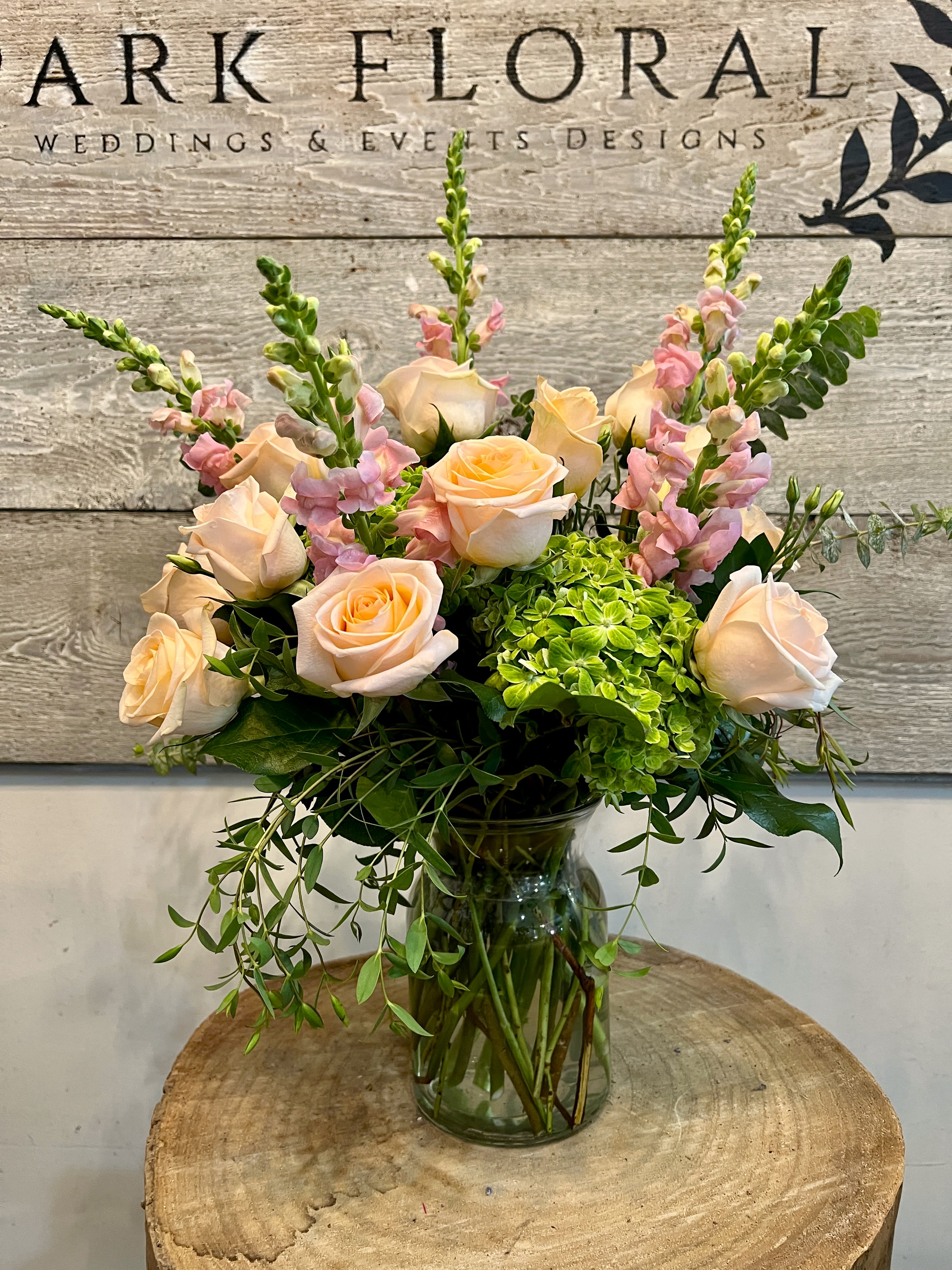 Peach Dreams  - Soft peach roses and pink snapdragons soften into a bright green hydrangea cluster for a garden-fresh look that reads romantic and effortlessly cheerful - perfect as a birthday bouquet, anniversary arrangement, or a thoughtful thank-you gift. This flower arrangement places the product type and occasion right up front so the person you're thinking of will immediately see the sentiment: warmth, appreciation, and a light, airy charm.  The design features layered, rounded styling with taller snapdragon spikes rising above the center and trailing greenery spilling gently from a clear glass vase. Color notes include peach, pink, white accents, and vivid green; flowers are primarily roses, snapdragons, and hydrangeas, arranged to create soft texture and visual depth. The clear glass vessel keeps the look natural and unfussy while allowing the blooms to be the focal point.  Made to feel like a freshly picked posy from a cottage garden, this arrangement suits recipients who appreciate soft color palettes and classic florals - a neighbor, mother, close friend, or colleague celebrating a milestone or simply needing a bright moment. Vases and stems arrive ready to display; refresh water every couple of days and recut stems if you want to extend bloom life.