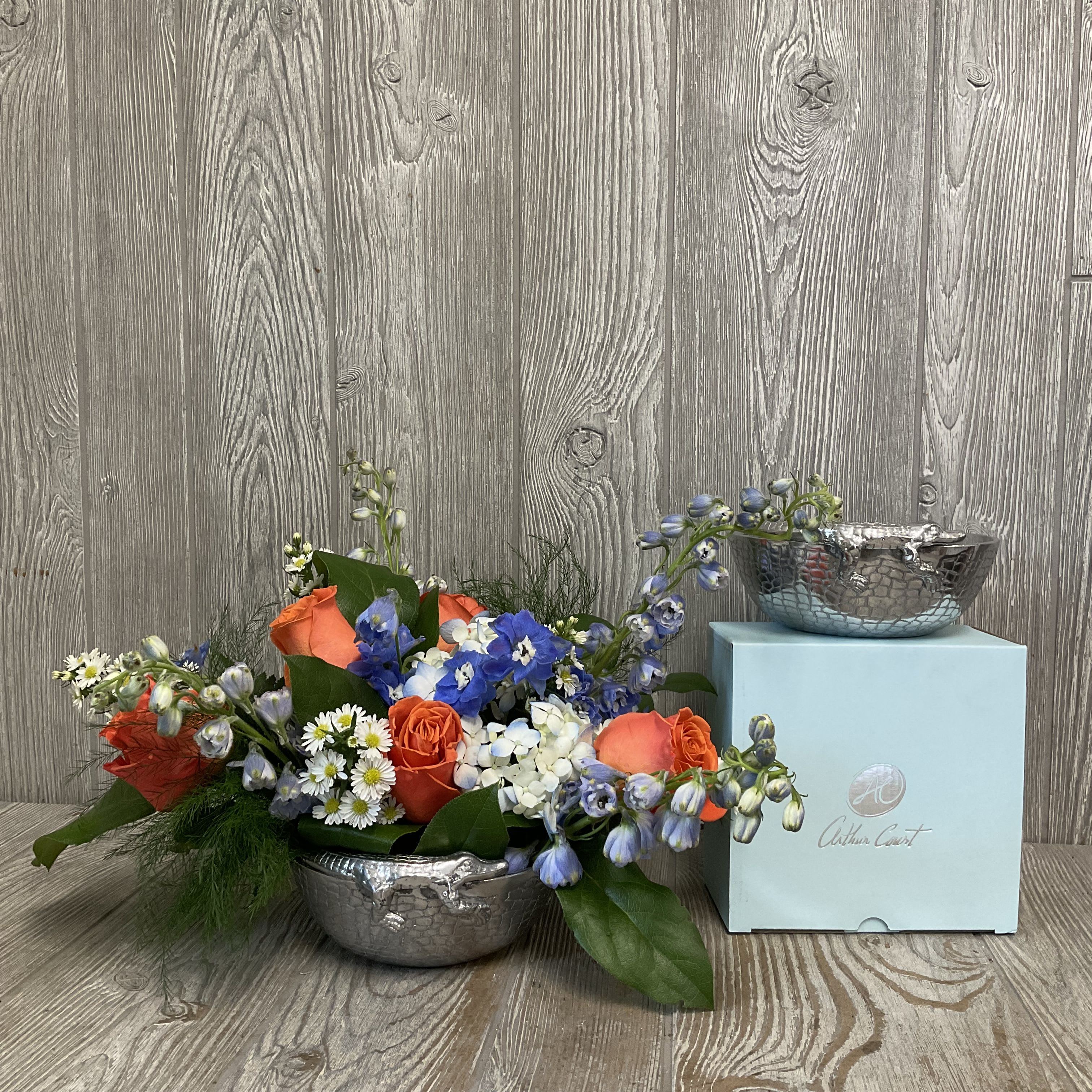 Gator Centerpiece by Arthur Court   - A playful hammered pewter bowl with an embossed alligator crawling along the rim holds a low, elongated floral design that instantly reads as spirited and refined - perfect as a graduation gift for the Gator grad, a bright birthday surprise, or a cheeky Father's Day or anniversary centerpiece. This flower arrangement pairs vibrant orange roses with tall, vivid blue delphinium, fluffy blue hydrangea and cheerful white monti to create an energetic, balanced palette that will make the recipient feel noticed and celebrated.  The arrangement sits in a reflective, reptile-textured metal bowl (approx. 6" Arthur Court keepsake) that doubles as a decorative accent after the flowers fade; the set is shown atop a light blue gift box for elegant presentation. The low, table-friendly shape works beautifully on dining tables, reception desks, or mantel displays and the mix of textures - silky roses, feathery delphinium, and cloud-like hydrangea - gives the composition both structure and softness.  Ideal for graduations, housewarmings, anniversaries, birthdays, and anytime you want to say "congrats" with personality, this keepsake-ready arrangement offers easy care: refresh water daily. It's the kind of gift that leaves the person you're thinking of feeling celebrated and seen - and leaves behind a stylish bowl they'll enjoy for years.