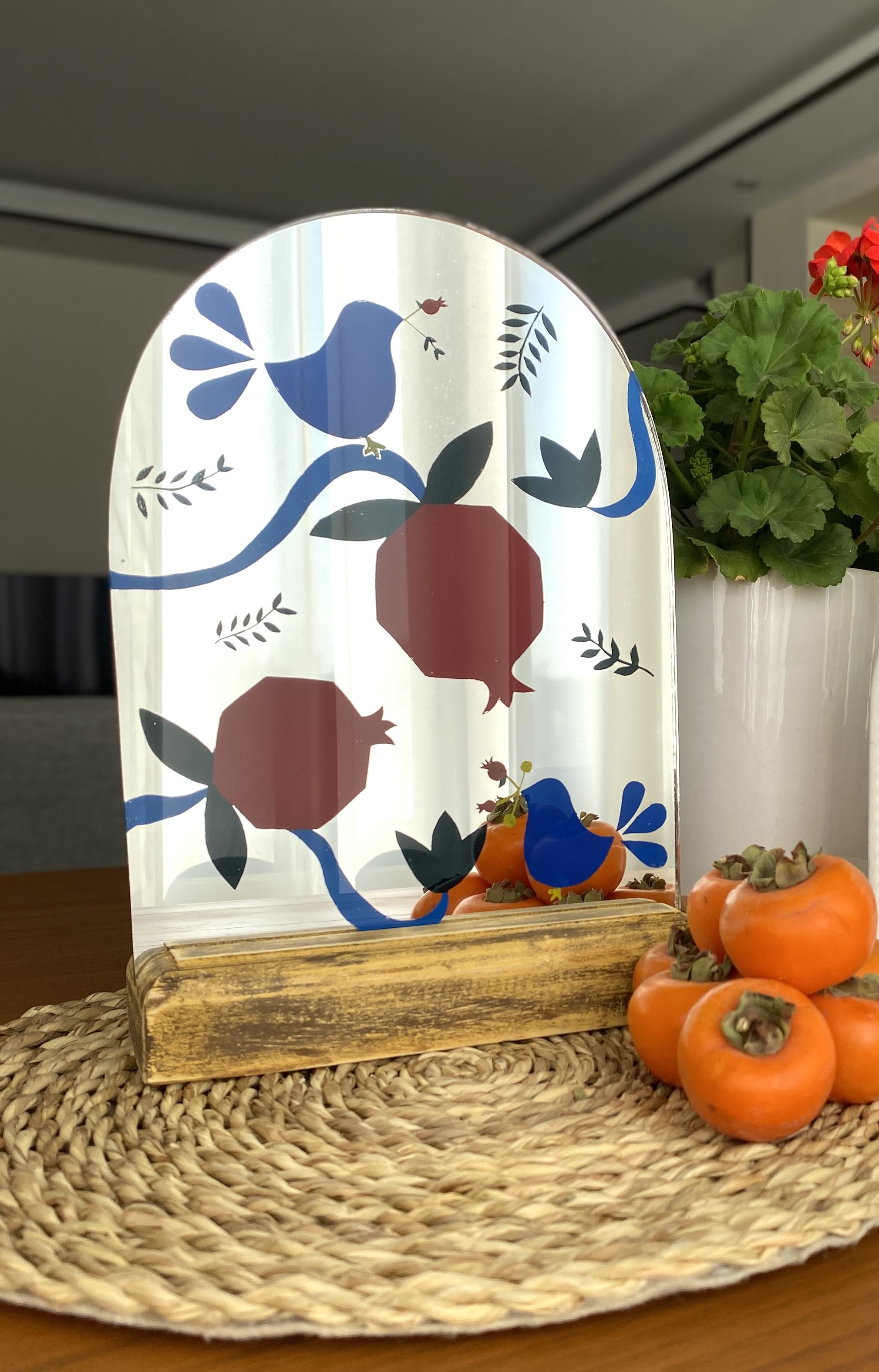 Decorative mirror with bird and pomegranate pattern beside potted flowers and persimmons