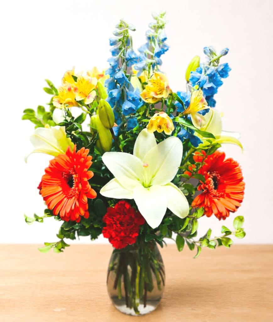Mixed bouquet with white lilies, orange gerbera daisies, blue delphinium, and red carnations in a glass vase