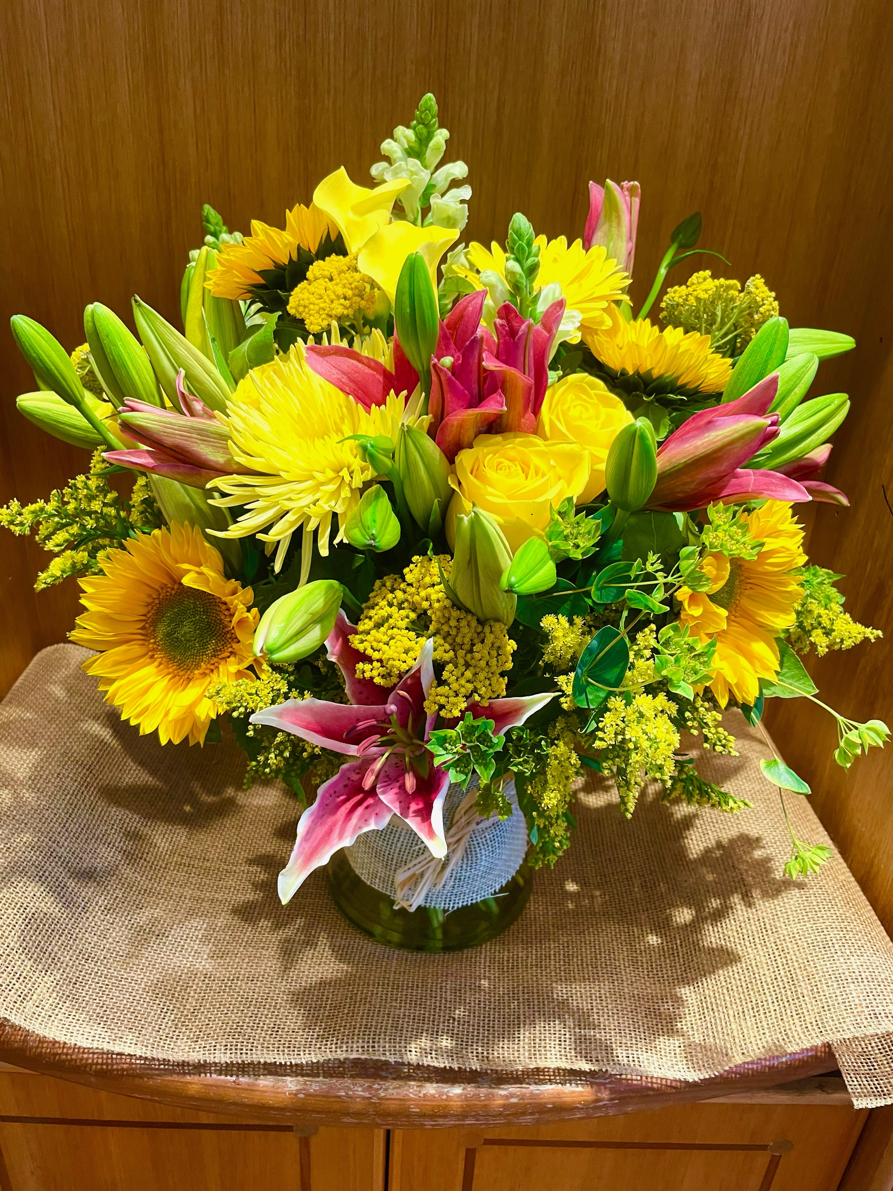 Mixed bouquet with yellow sunflowers, roses, lilies, and pink blooms in a glass vase