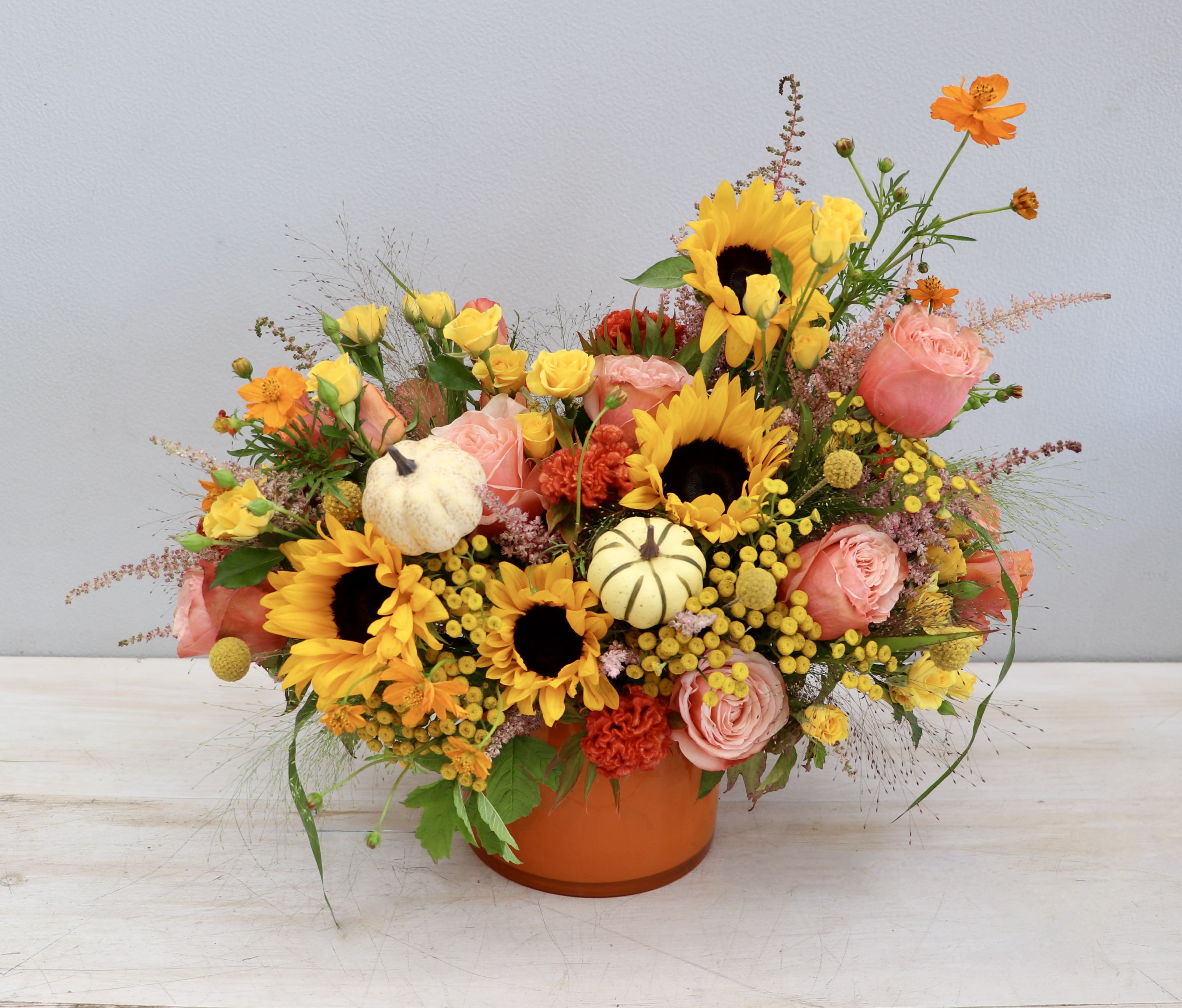 Low orange pot arrangement with sunflowers, peach roses, yellow blooms and small white pumpkins.