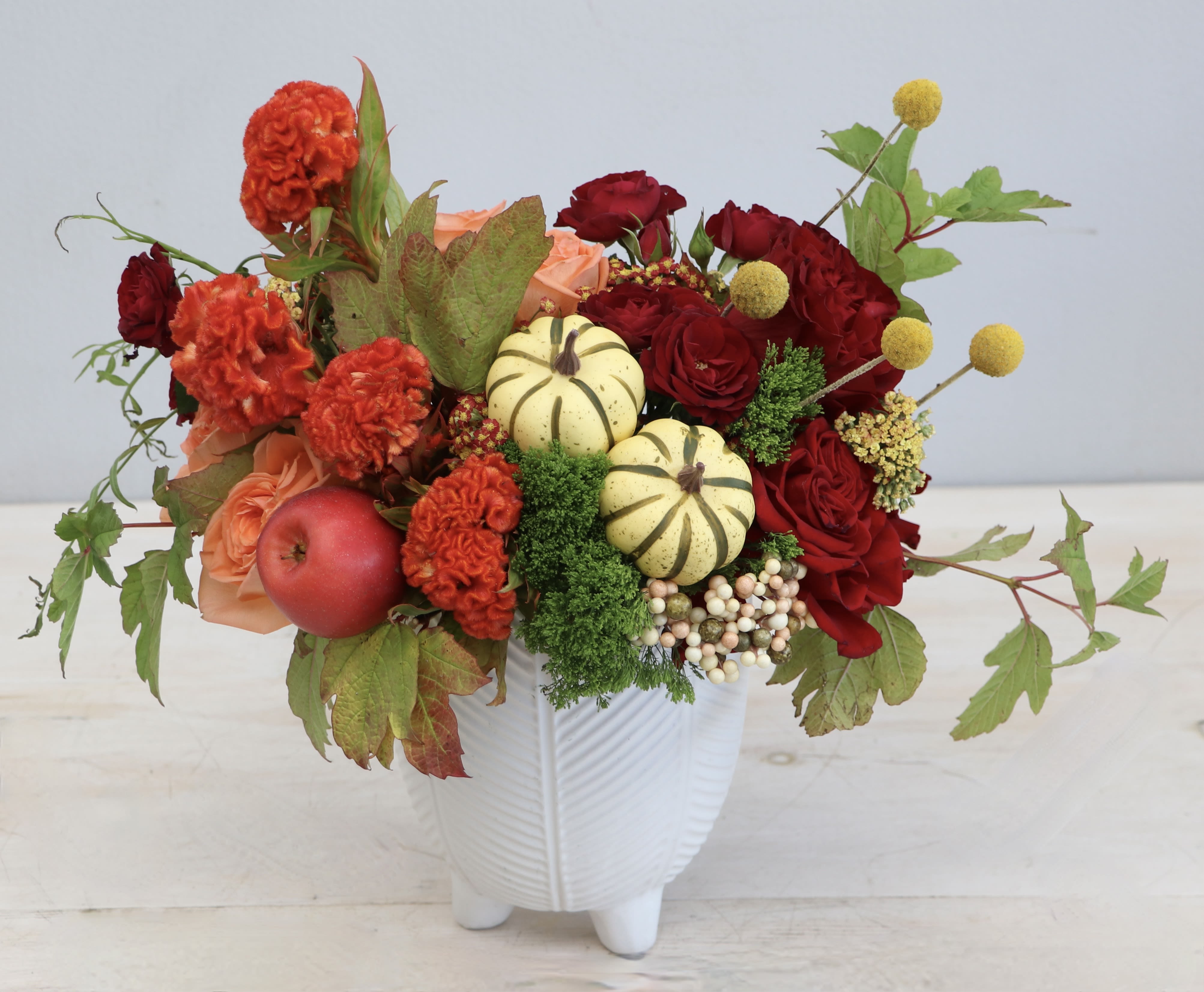 Autumn arrangement with red and orange flowers, mini pumpkins, and an apple in a white ceramic vase