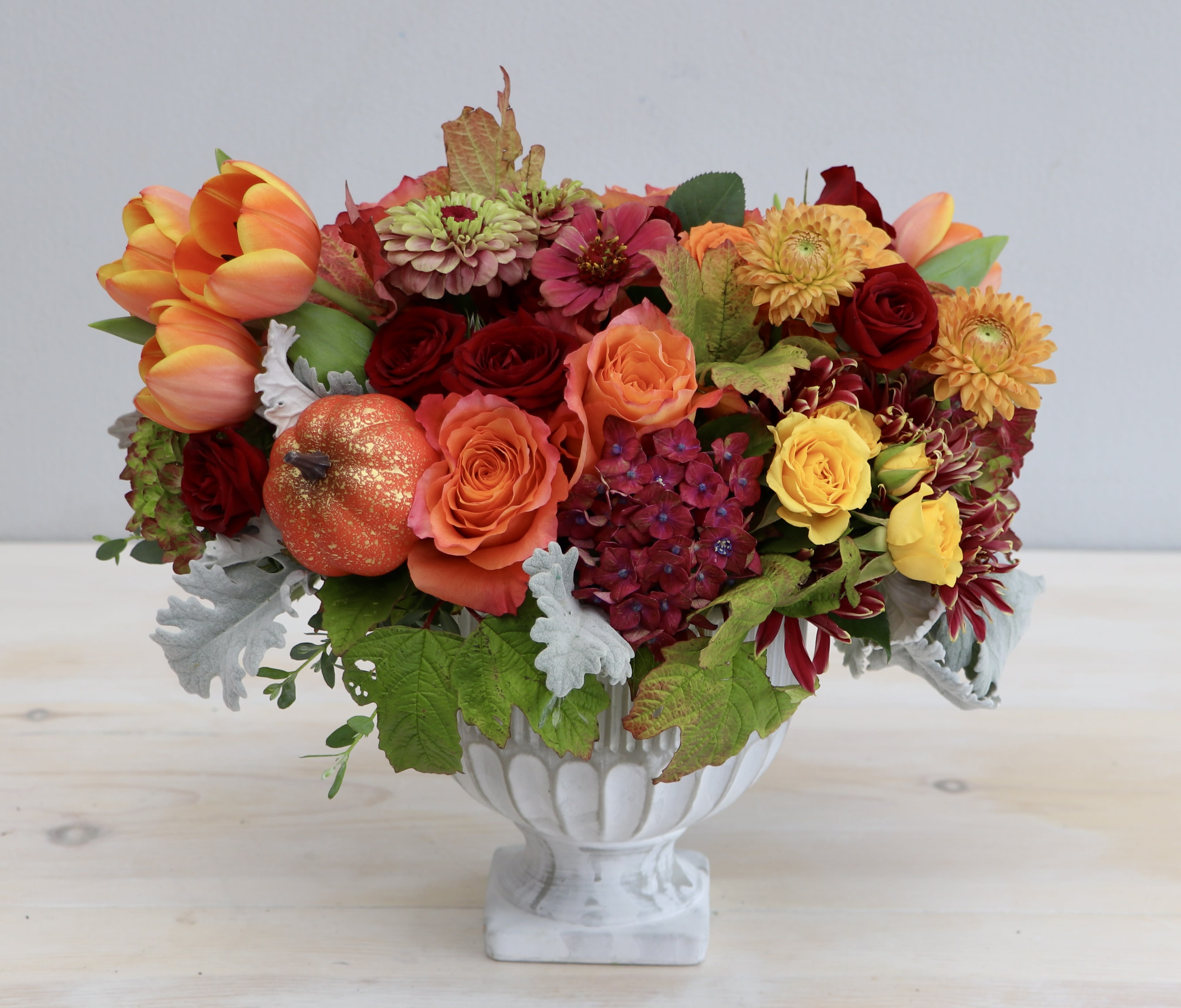 Low orange and red flower arrangement with roses and tulips in a white pedestal bowl.