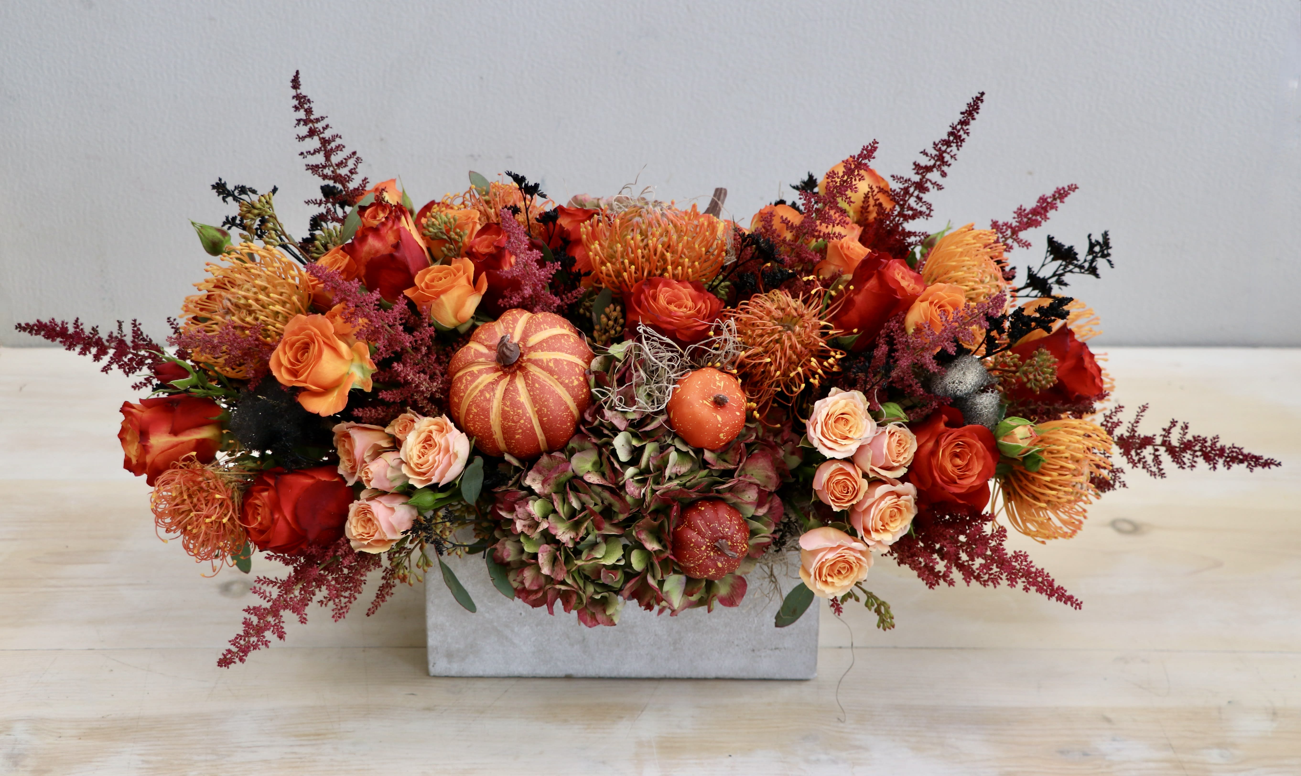 Low rectangular centerpiece with orange and red roses, protea, hydrangea, and small pumpkins.