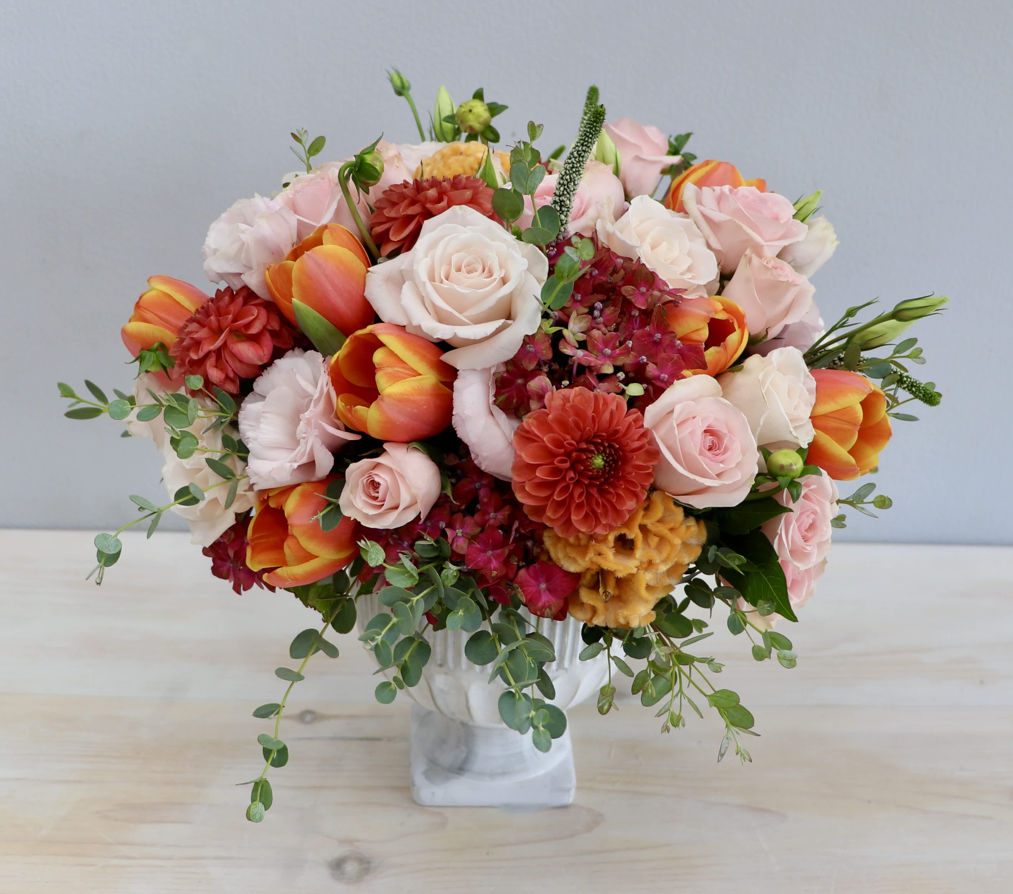 Rounded arrangement of pink roses, orange tulips, and mixed blooms in a white pedestal vase.