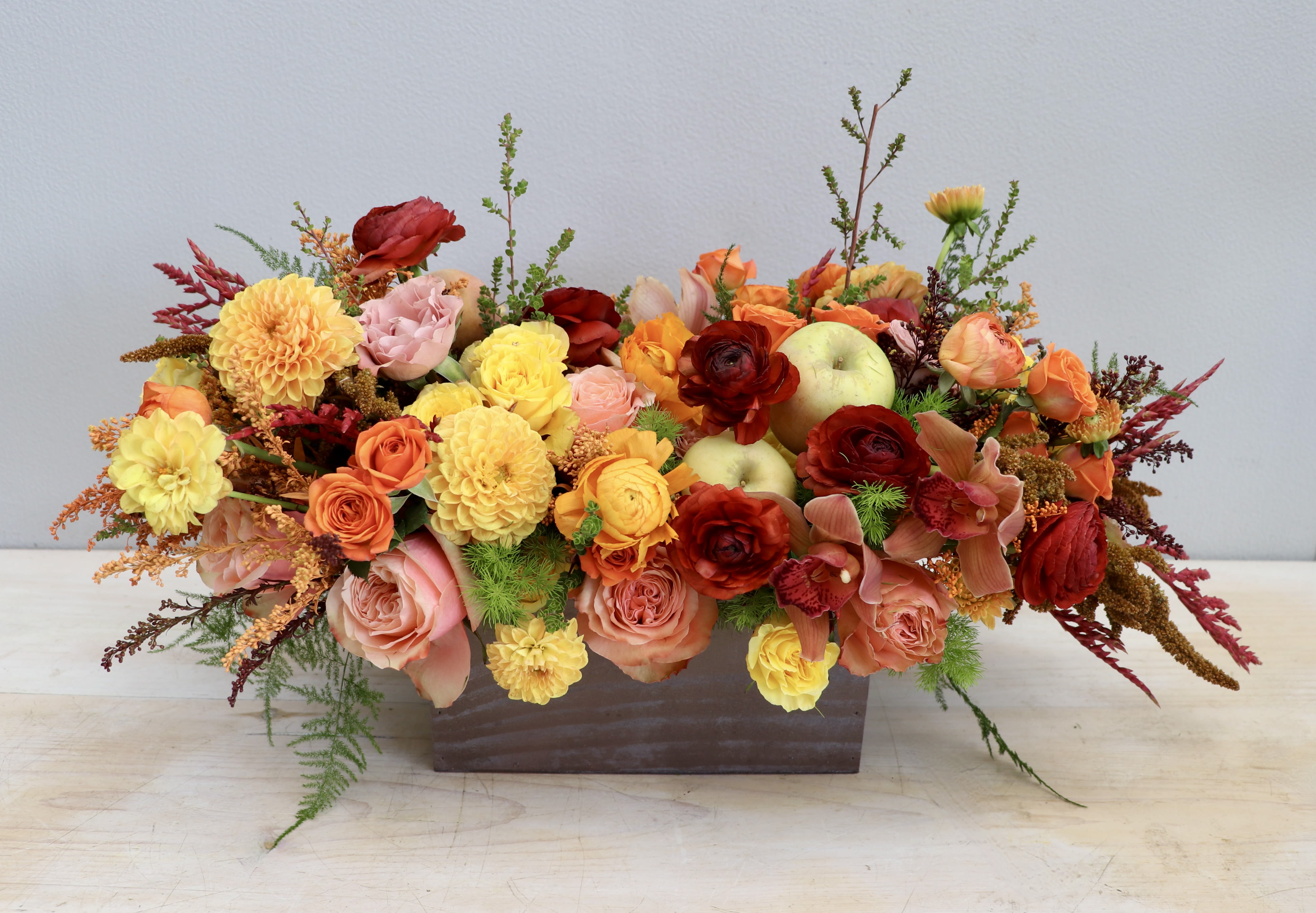 Low rectangular arrangement of orange, yellow, and red flowers with apples in a metal container
