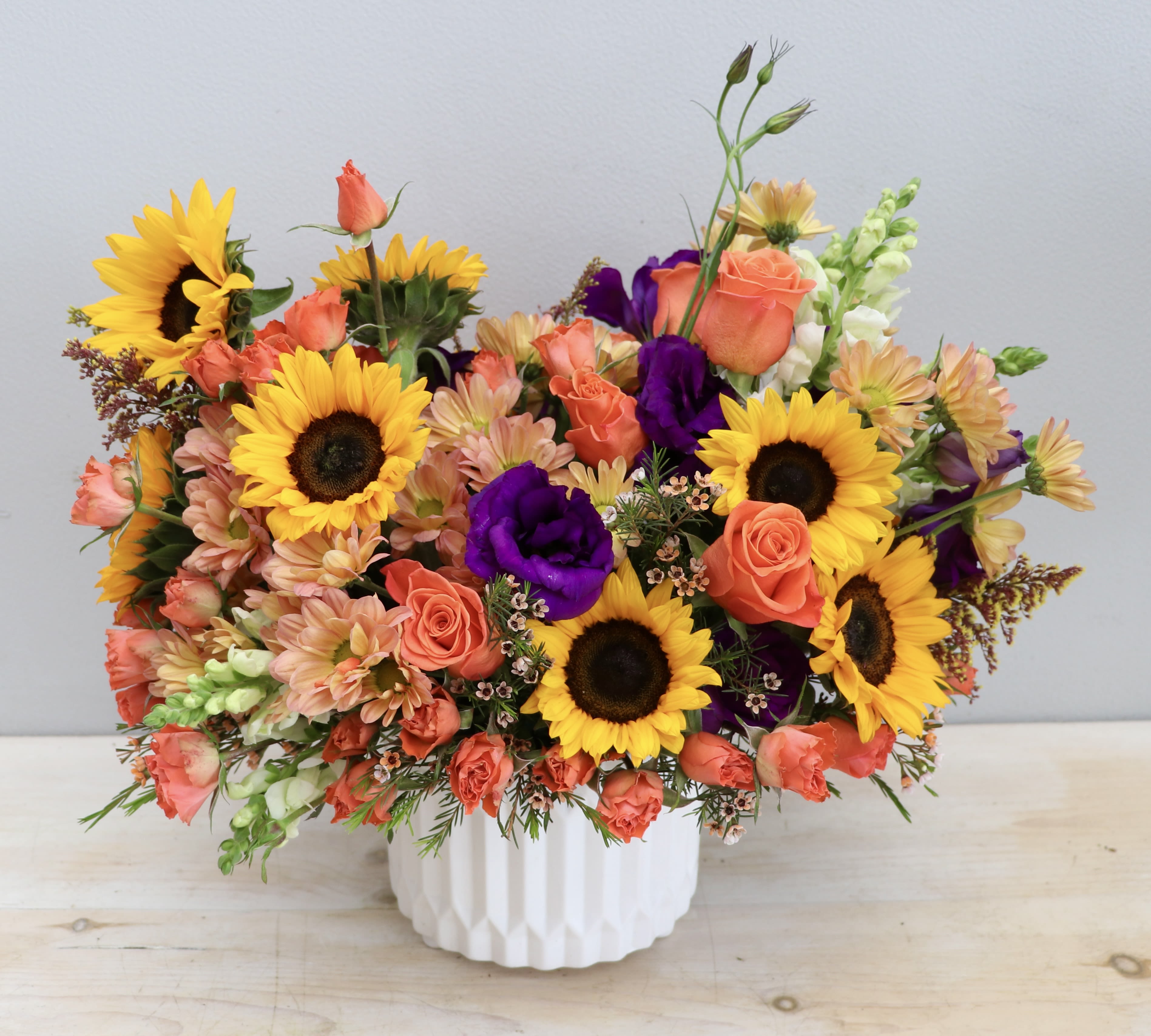 Bright arrangement of sunflowers, orange roses, and purple blooms in a white ceramic vase