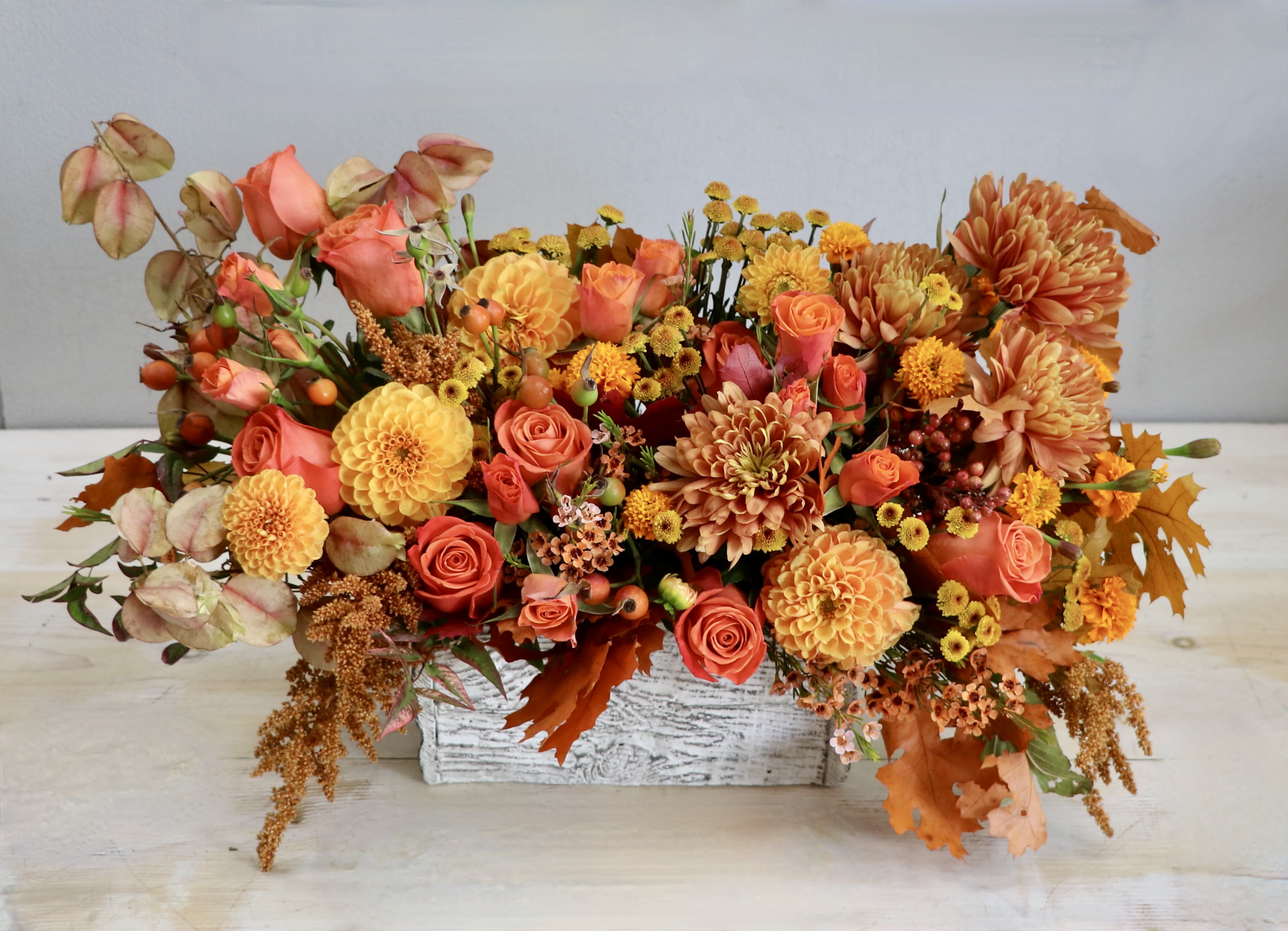 Low rectangular arrangement of orange and yellow roses, dahlias and mums in a white wood box.