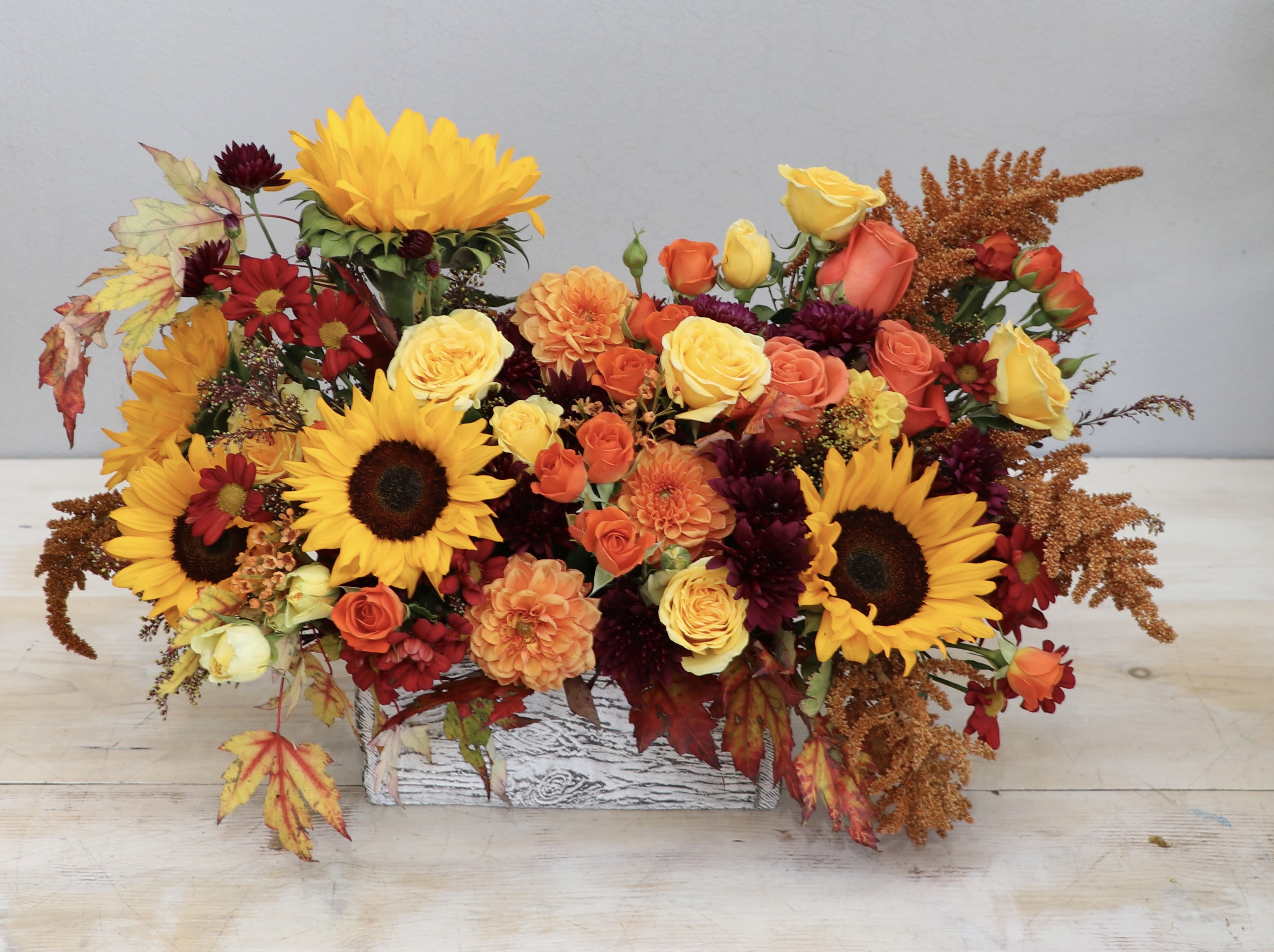 Low rectangular arrangement of sunflowers, roses, and fall-toned blooms in a whitewashed wooden box.