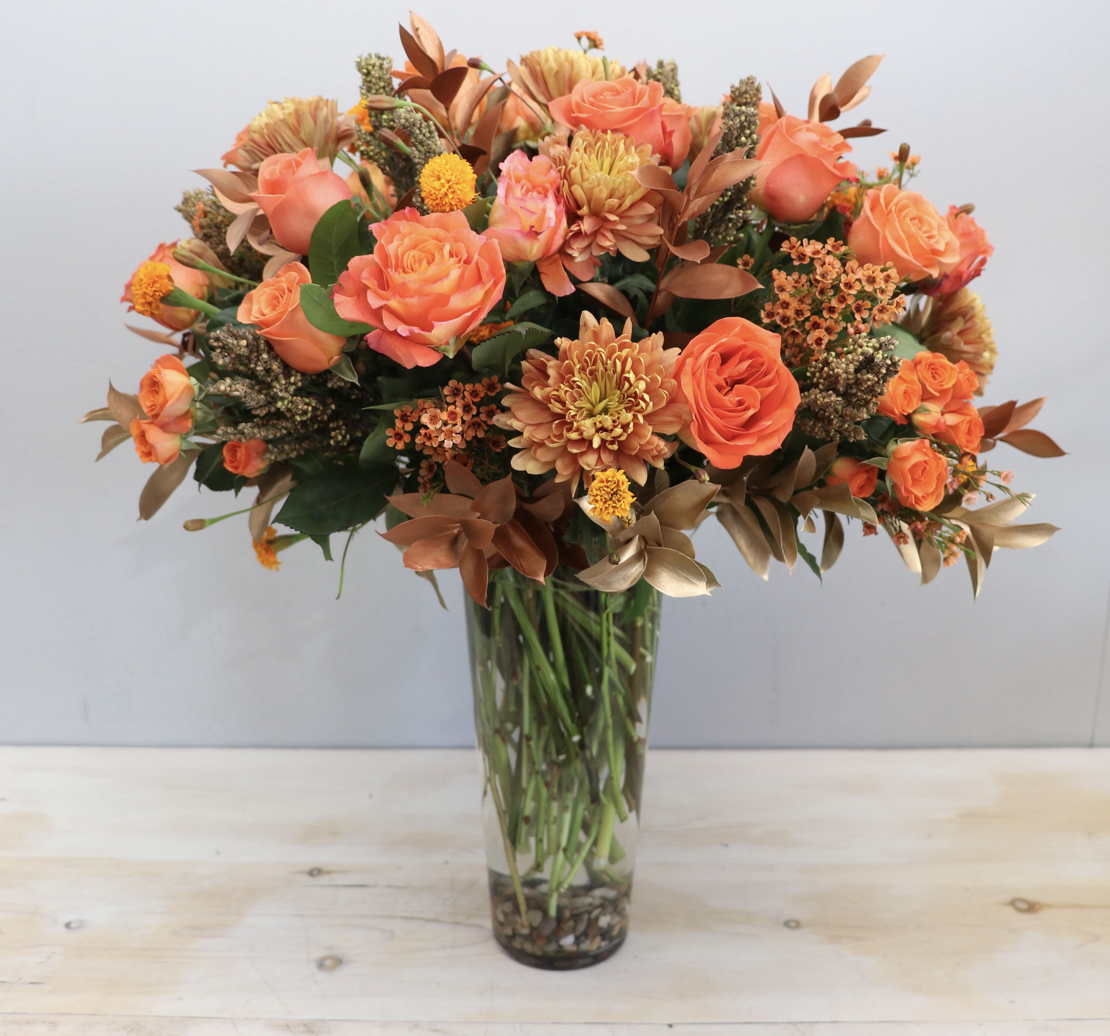 Tall vase arrangement of orange roses and chrysanthemums with bronze accents in a clear glass cylinder