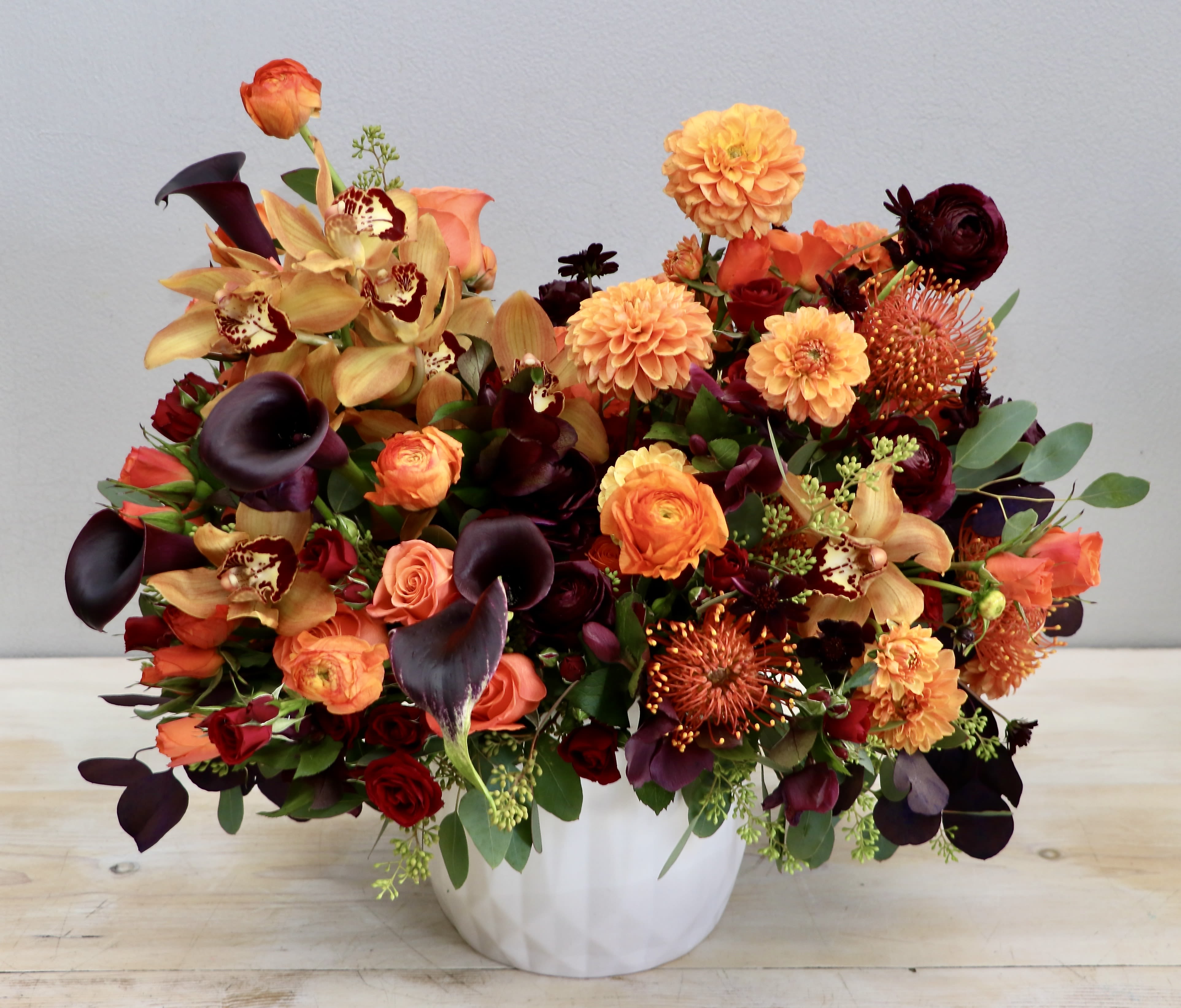 Low white vase filled with orange and burgundy flowers including roses, orchids, dahlias, and calla lilies