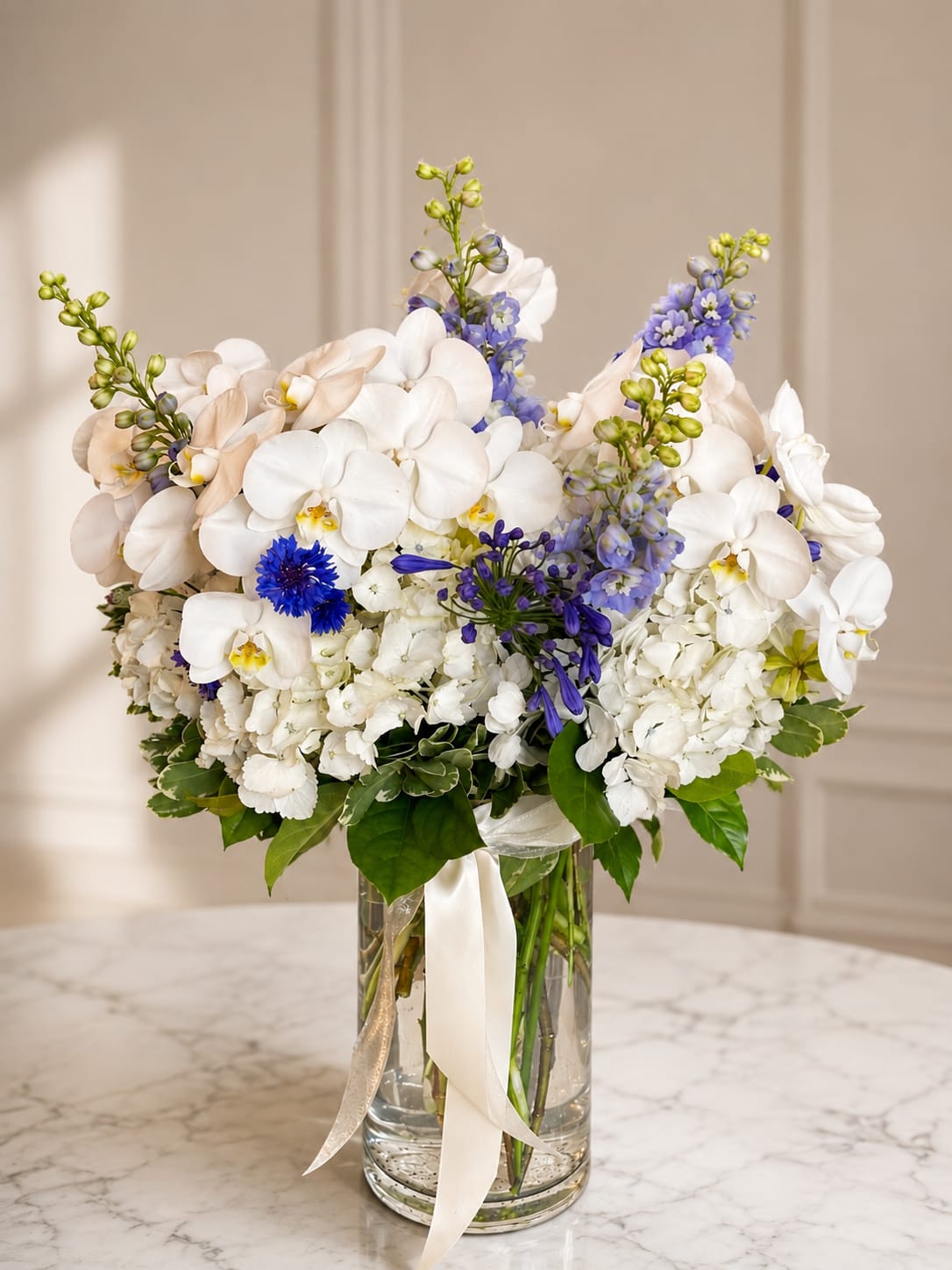 White orchids and blue flowers arranged in a clear glass vase with ribbon
