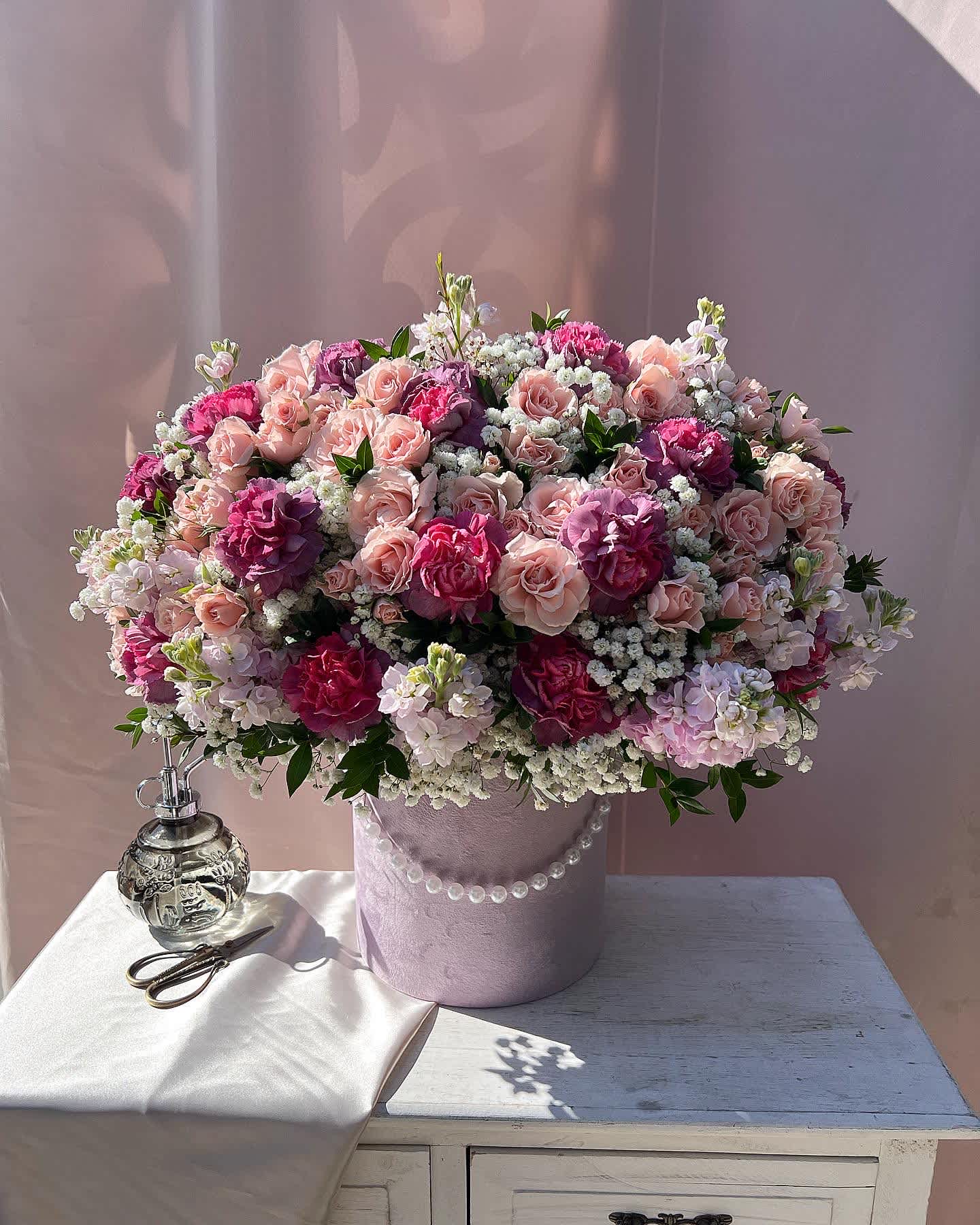 Pink Mood Box D - DELUXE size is shown in the picture.  This charming arrangement showcases a delightful mix of vibrant carnations, delicate spray roses, and fragrant stock, all beautifully complemented by airy white gypsophila. The combination of bold and soft blooms creates a balanced and textured display, perfect for brightening any space or celebrating special moments. Its cheerful aesthetic makes it an ideal choice for gifts, events, or simply adding a touch of joy to your home. For arrangements in boxes and ceramic pots, Glass Vases are NOT needed.