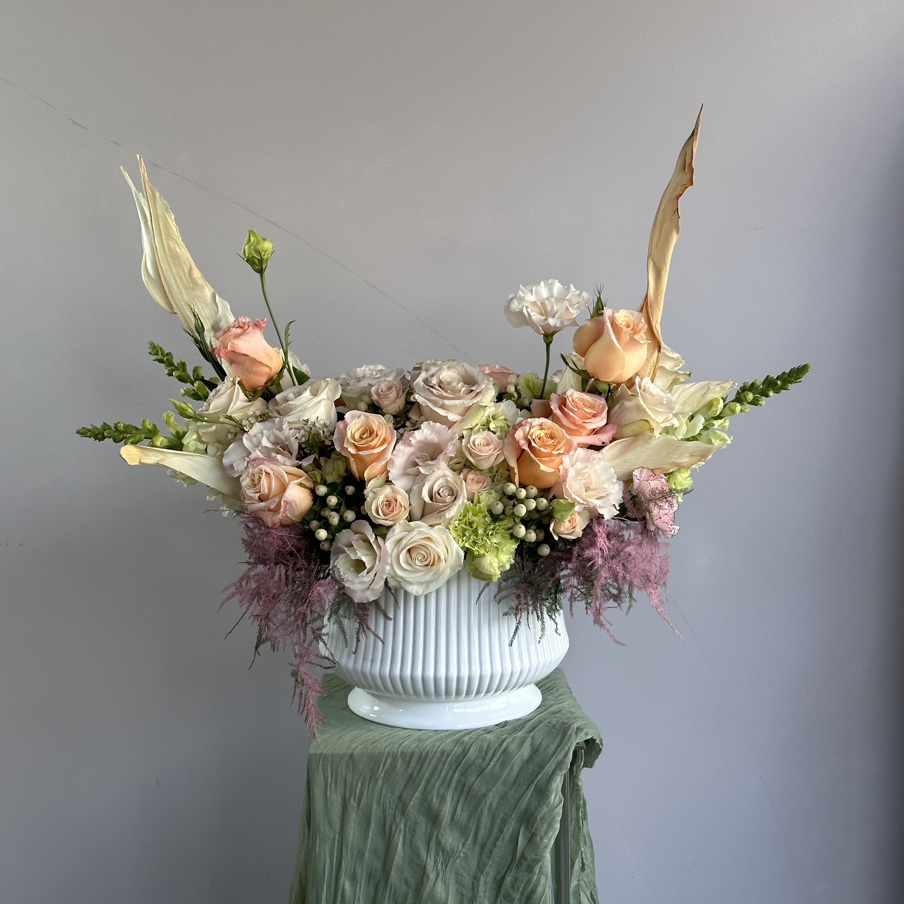 Sofi Vase P - This elegant floral arrangement features soft peach and blush roses, complemented by delicate white blooms and vibrant green accents. The mix of textures, including unique pink feathers, adds a modern and luxurious touch, perfect for a sophisticated centerpiece or event decor. Placed in a sleek, white vase, it's a timeless display of beauty and grace. PREMIUM size is shown in the picture