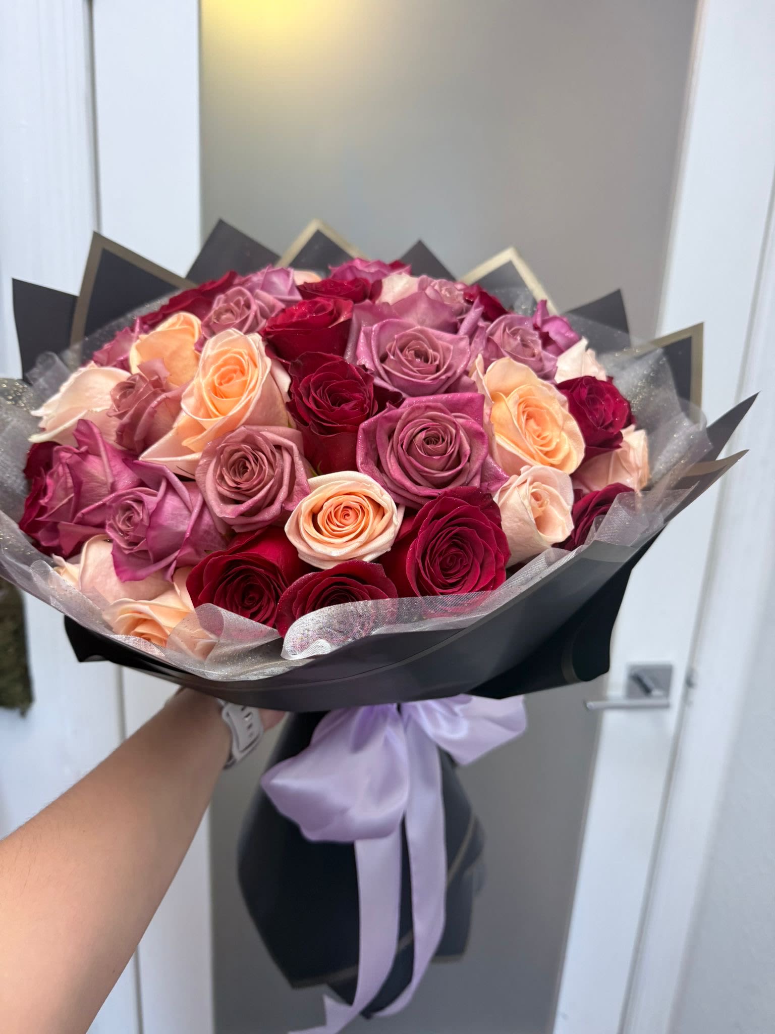Bouquet of pink, peach, and red roses wrapped in black paper with a lavender ribbon