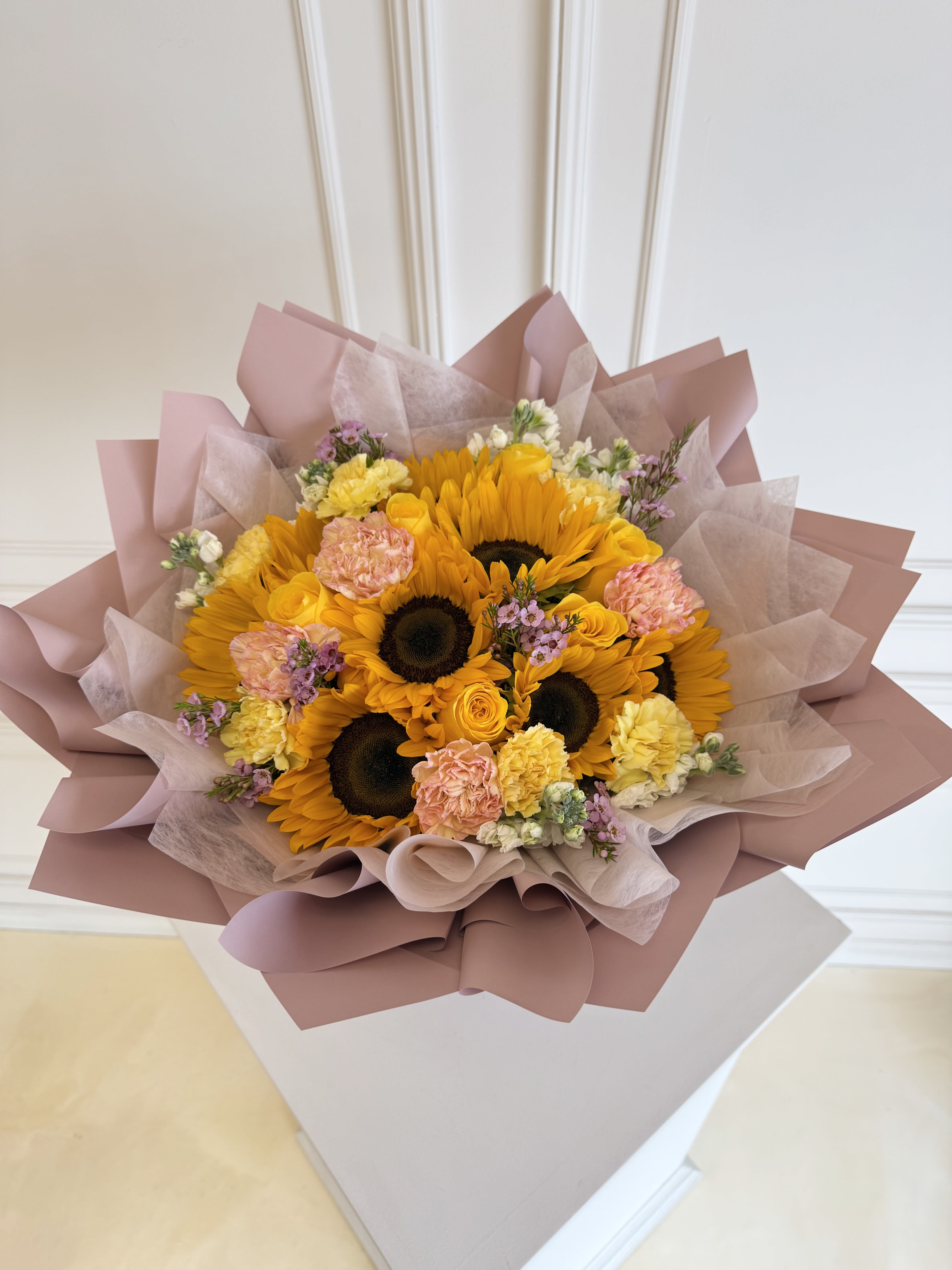 Bouquet of sunflowers, yellow roses, and pink carnations wrapped in blush paper