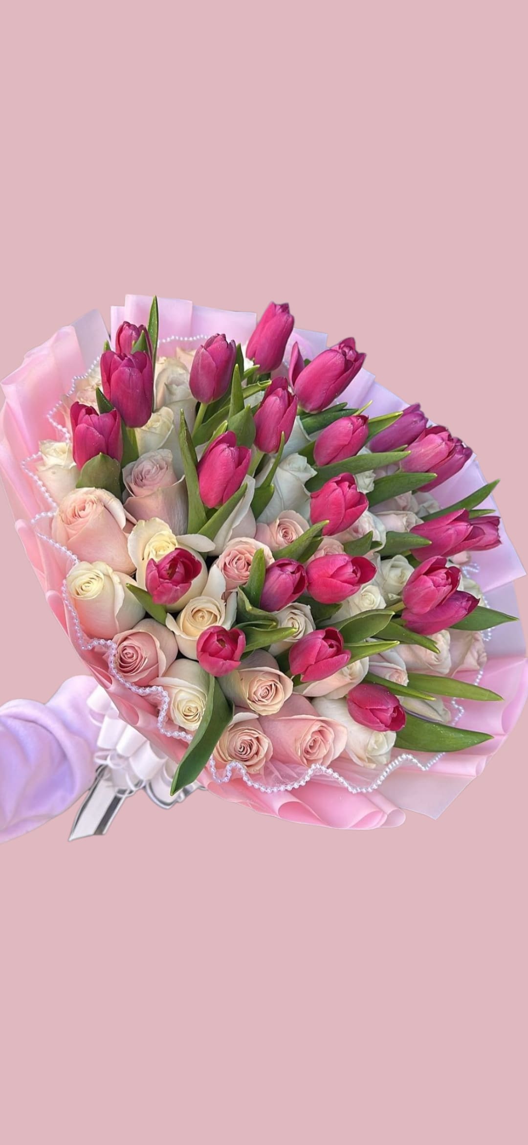 Sweet Tulip Bouquet - A lively cluster of hot pink tulips and soft cream-to-blush roses creates an energetic yet romantic birthday bouquet or anniversary arrangement that will make the person you're thinking of feel celebrated. The bright tulips rise above the rounded roses for a pleasing contrast in height and color, delivering a joyful, feminine impression perfect for romantic gestures, congratulations, or a thoughtful pick-me-up.  Carefully wrapped in layered pink paper with a scalloped, beaded trim and tied with a white ribbon, this arrangement looks polished straight out of the wrap-no extra fuss required. Colors: pink, white, green. Ideal for giving as-is; for freshness, trim stems and change the water every other day to help blooms last longer.