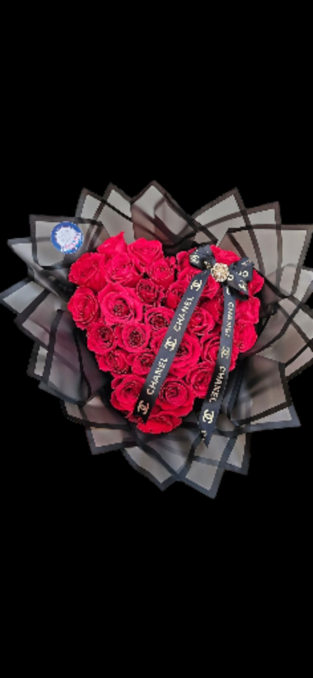 Heart bouquet  - A dense heart-shaped cluster of bright red roses makes an unmistakable statement-the perfect romantic bouquet for an anniversary, Valentine's Day, or any moment you want to declare your affection. The heart motif and high-contrast red-and-black palette create a dramatic, couture look that will make the person you're thinking of feel deeply seen and celebrated.  Each rose is arranged tightly to form a solid red heart and is framed by multiple layers of structured black translucent wrapping that fan outward like a sculptural halo. A black logo ribbon with white text is tied across the blooms and finished with a jeweled brooch and round badge for a luxurious, fashion-forward accent. This arrangement reads as both classic and modern-bold color, refined details, and tactile textures that catch the eye from every angle.  Ideal for lovers, milestone celebrations, or a striking sympathy of devotion, this bouquet arrives ready to display; no extra vase required. The structured wrapping helps protect the blooms in transit-order with confidence to deliver a powerful, memorable message.