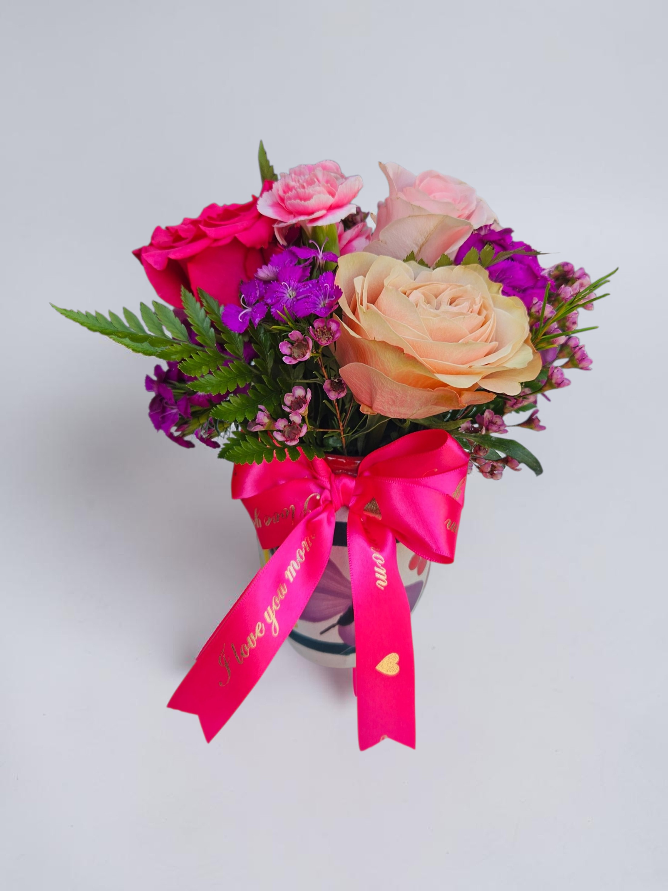 Cantarito de Amor - A joyful cluster of roses and carnations in soft peach, pale pink, hot pink and purple makes this flower arrangement an irresistible birthday bouquet or anniversary gift for the person you want to make feel truly cherished. The compact, rounded design reads as feminine and festive-perfect for Valentine's Day, Mother's Day, or a just‑because surprise that says "you matter" in bright, romantic color.  Roses take center stage-large peach and pale pink blooms paired with vivid hot‑pink roses-balanced by carnations and small clustered purple filler flowers for texture and depth. Fern‑like foliage frames the composition, adding airy green contrast, while a bright pink satin ribbon finishes the look. Choose the hand‑painted ceramic vase with a colorful butterfly motif for a decorative keepsake, or the clear glass vase tied with pink ribbon for a classic, modern display.  Sized to fit a tabletop or bedside, this arrangement arrives ready to gift and brightens any small space; it's a sweet, everyday expression of affection that will make her feel genuinely celebrated.
