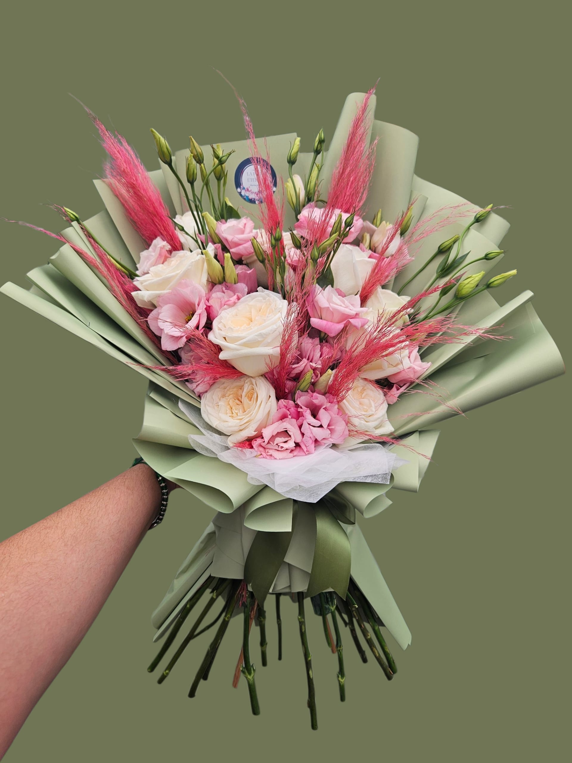 Elegant Mom  - Blush pink and creamy white roses mingle with soft pink lisianthus and bright feathery pink accents to create a romantic, airy flower arrangement perfect as a Mother's Day bouquet, anniversary roses, or a cheerful birthday gift. The hand-tied composition reads fresh and feminine-she'll feel truly seen when presented with the soft pastels and playful texture that lift this bouquet above everyday flowers.  This arrangement features cream and blush roses, pink lisianthus, upright green lisianthus buds for movement, and bright pink feathery grass for height and texture. Wrapped in pale green decorative paper with tissue and bouquet wrap, it's finished with white tulle ribbon and a dark green satin ribbon for a polished, garden-style look. The result is a refined yet whimsical bouquet in pink, white, green, and peach tones that arrives ready to display or carry.  Ideal for birthdays, anniversaries, thank-you gestures, or a thoughtful just-because surprise, this arrangement offers a soft, celebratory mood without being overdone. Choose your size at checkout for the perfect impact; stems are arranged hand-tied for natural movement-replace water daily and trim stems to extend the life of the blooms.