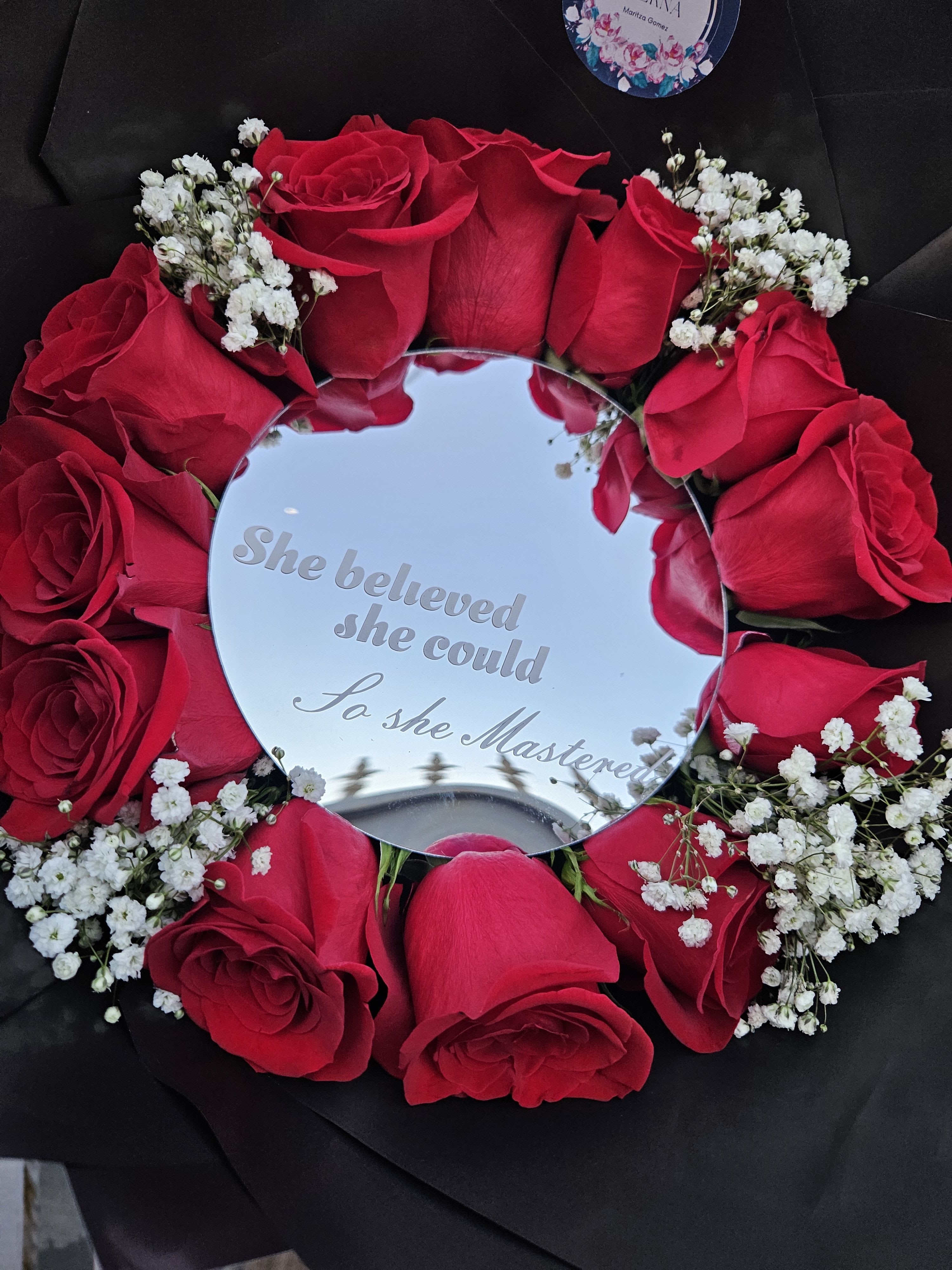 She Believed Graduation Bouquet  - A bold circle of deep red roses arranged around a mirrored center creates an unforgettable anniversary or Valentine's Day flower arrangement that reads as both romantic gift and keepsake. The mirrored plaque at the heart of the design carries engraved text and reflects the moment, so the person you're thinking of will see themselves in a thoughtful, commemorative presentation-perfect for marking an anniversary, a special date, or a heartfelt declaration of love.  The roses are packed tightly in a round formation with clusters of small white filler flowers threaded between the blooms for delicate contrast and texture. Black wrapping paper frames the arrangement, heightening the red-and-white palette, while a bright red ribbon and a round decorative pick finish the dramatic look. The piece arrives in a clear plastic sleeve to protect the petals and the engraved mirror; the compact, circular silhouette makes it easy to display as a tabletop tribute or a keepsake centerpiece that feels both intimate and celebratory.