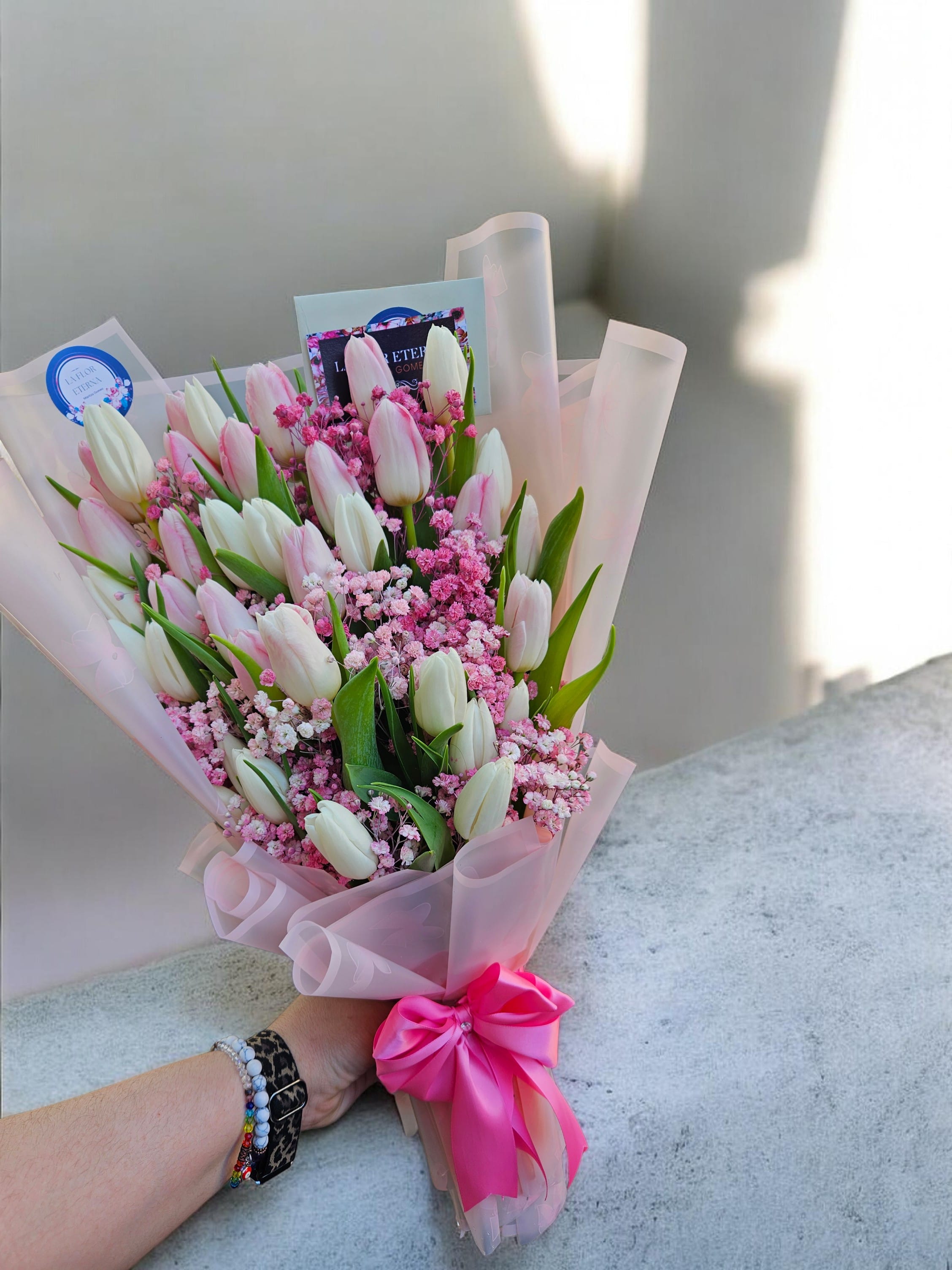 Pink Paradise - Soft pale pink and creamy white tulips gathered with tiny pink filler flowers create an immediate feeling of gentle celebration - perfect as a Mother's Day bouquet or a birthday, anniversary, or "thinking of you" surprise. This compact, upright hand-tied arrangement reads romantic without being over-the-top, making it an excellent choice for love and romance, new baby visits, or a sweet "I'm sorry" gesture that feels sincere.  The arrangement pairs those tulips and delicate fillers with translucent blush-pink wrapping paper and a bright pink ribbon bow for a light, feminine look. A greeting card is tucked into the top and a round label sticker finishes the package, so it's ready to hand to the person you're celebrating. Its compact size makes it easy to carry to a workplace, bedside, or small-table display.  Send this bouquet as a thankful, congratulatory, or comforting note - from retirements and graduations to get-well wishes and just-because moments - and she'll feel genuinely seen. For best enjoyment, trim stems and refresh water every couple of days to keep the tulips perky; if you need it delivered same-day or scheduled for a holiday like Valentine's Day or Women's Day, choose your delivery date at checkout.