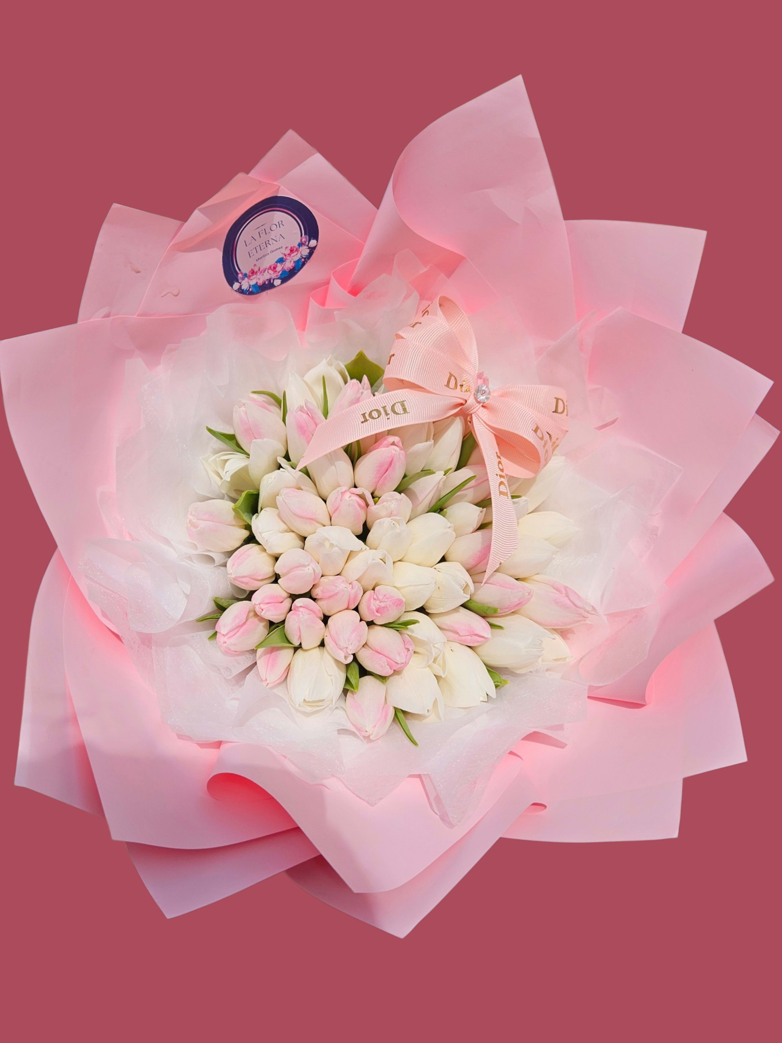 Dreamy Tulips - A rounded dome of white tulip buds kissed with soft pink tips makes a quietly romantic statement - perfect as a birthday bouquet, anniversary gift, or a "just because" surprise for someone you adore. The arrangement reads feminine and affectionate at first glance, the pale palette and compact shape saying thoughtful and intimate rather than extravagant.  Each tulip cluster is wrapped in translucent white paper layered with wide pink decorative paper that fans out like extra petals, then finished with a pale pink fabric ribbon bow and a round branded label for an elegant touch. The buds are tightly arranged for a full, textured look; curling white ribbons from the included red, pink, and white glossy latex balloons add a playful, celebratory lift when ordered together, making the set ideal for parties or romantic gestures.  This is the kind of gift that makes the recipient feel genuinely seen-soft, fragrant tulips that read both tender and festive. Available in a compact, hand-held size; order for same-week delivery to ensure buds arrive fresh and gently closed so they open beautifully over the next few days.