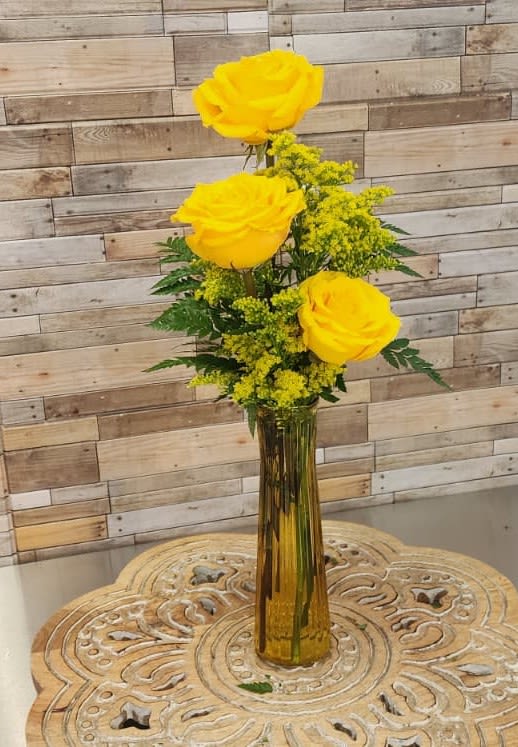 Tall amber vase with three yellow roses and yellow filler flowers on a carved wooden table.