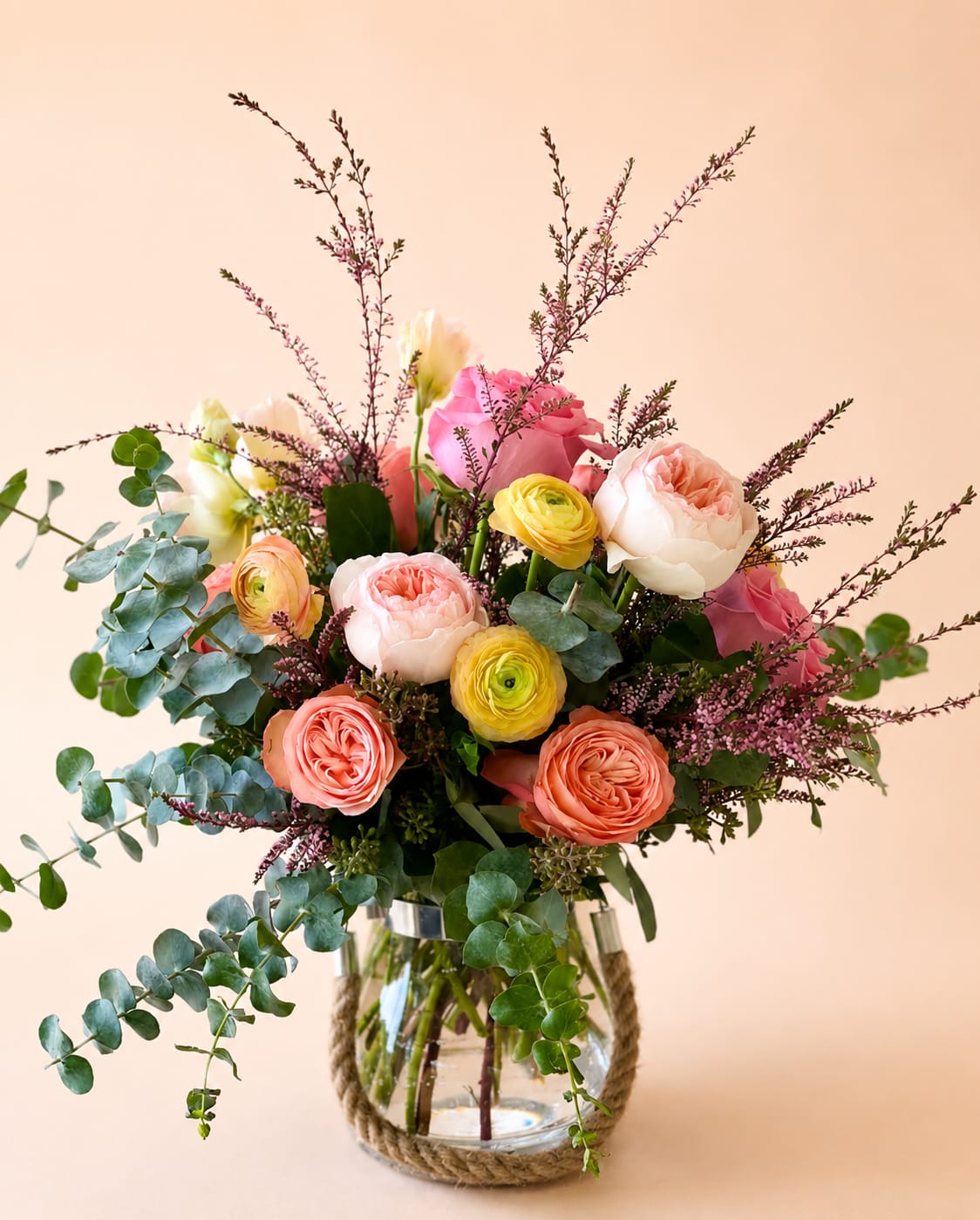 Fleurs du Ranch  - Our Arrangement "Fleurs du Ranch" features some of our most premium florals such as: Kahala Garden Roses, Peach Osteen Garden Roses, Saga Garden Roses, Expression Garden Roses, Pink Scottish Heathers, Sunshine Ranunculus, Light Blush Lisanthus, and Spiral Eucalyptus built inside a thick glass rope vase. Inspired by the rustic, organic,  Ranch lifestyle.