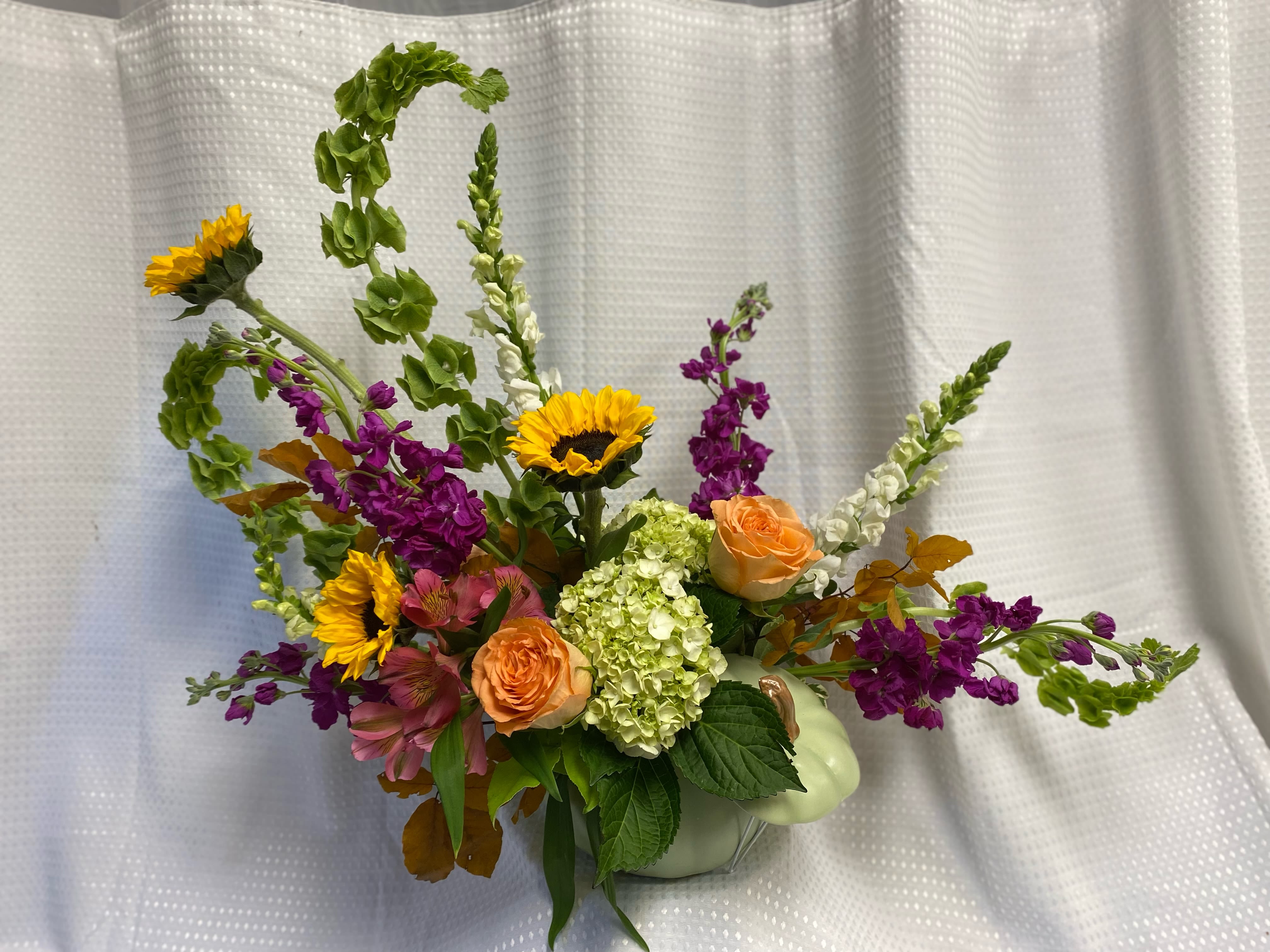 Mixed bouquet with sunflowers, peach roses, and purple blooms in a pale green pumpkin vase