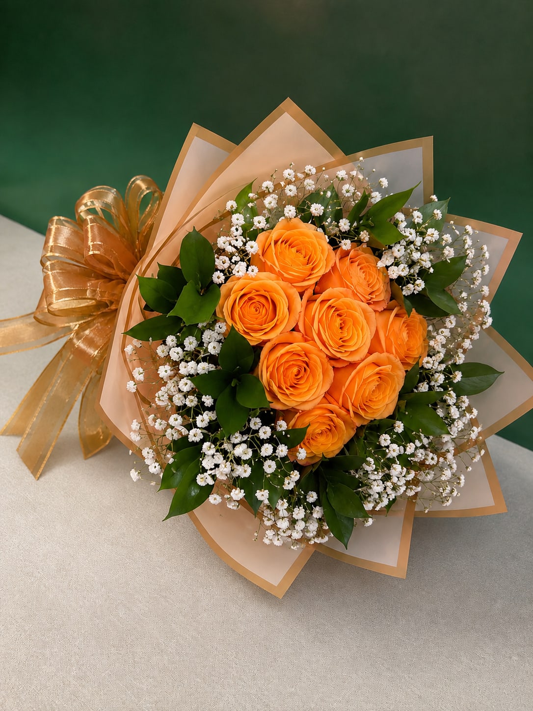 Citrus Orange Glow Bouquet - Tightly clustered bright orange roses form the heart of this Citrus Orange Glow bouquet, a joyful birthday and just-because flower arrangement that also reads beautifully for anniversaries, housewarmings, or a thoughtful I'm Sorry gesture. The warm orange and gold palette creates an immediate sense of cheer-she'll feel noticed and celebrated the moment the bouquet arrives.  The design pairs clustered orange roses with delicate white filler blooms and glossy green foliage for contrast and texture. Wrapped in layered peach and translucent decorative wrapping paper with gold edging and finished with a large metallic gold ribbon, the presentation is polished and gift-ready. Approximately eight citrus roses compose the focal cluster, surrounded by fresh greenery; substitutions may be made with flowers of equal or greater value while keeping the same style and color story.  Perfect for saying thank you, thinking of you, congrats, or a romantic note, this arrangement arrives ready to display and brightens any room. Care: keep stems trimmed, change water every other day, and place away from direct heat to prolong freshness. Delivered in New York, NY and surrounding areas.