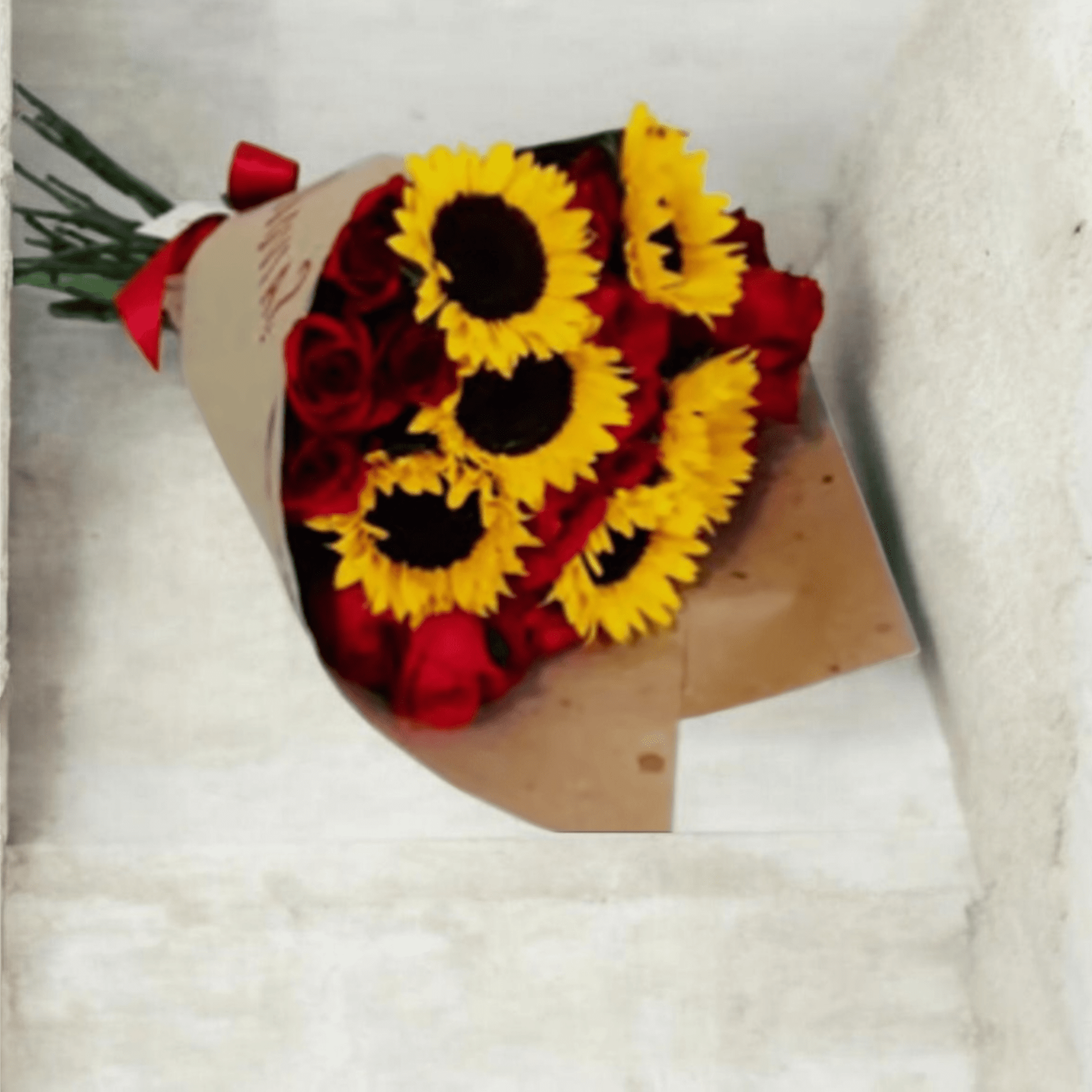 Simona Sunflowers and Red Roses Bouquet - Striking red roses paired with sunny yellow sunflowers create an unforgettable contrast in this hand-tied flower arrangement-perfect as an anniversary or birthday bouquet or a heartfelt "just because" surprise. The mix reads both romantic and joyful, making the person you're thinking of feel cherished and genuinely celebrated.  Tightly gathered and wrapped in rustic brown paper, SIMONA is finished with a red ribbon for a simple, modern presentation that lets the blooms take center stage. The bouquet combines deep, velvety red roses with several bright sunflowers for a warm, saturated palette that pops in any room.  Ideal for saying thank you, marking a milestone, or brightening an ordinary day, this arrangement balances classic romance with cheerful energy. Delivered ready to display, the blooms arrive hand-tied-just trim the stems, add fresh water, and enjoy.
