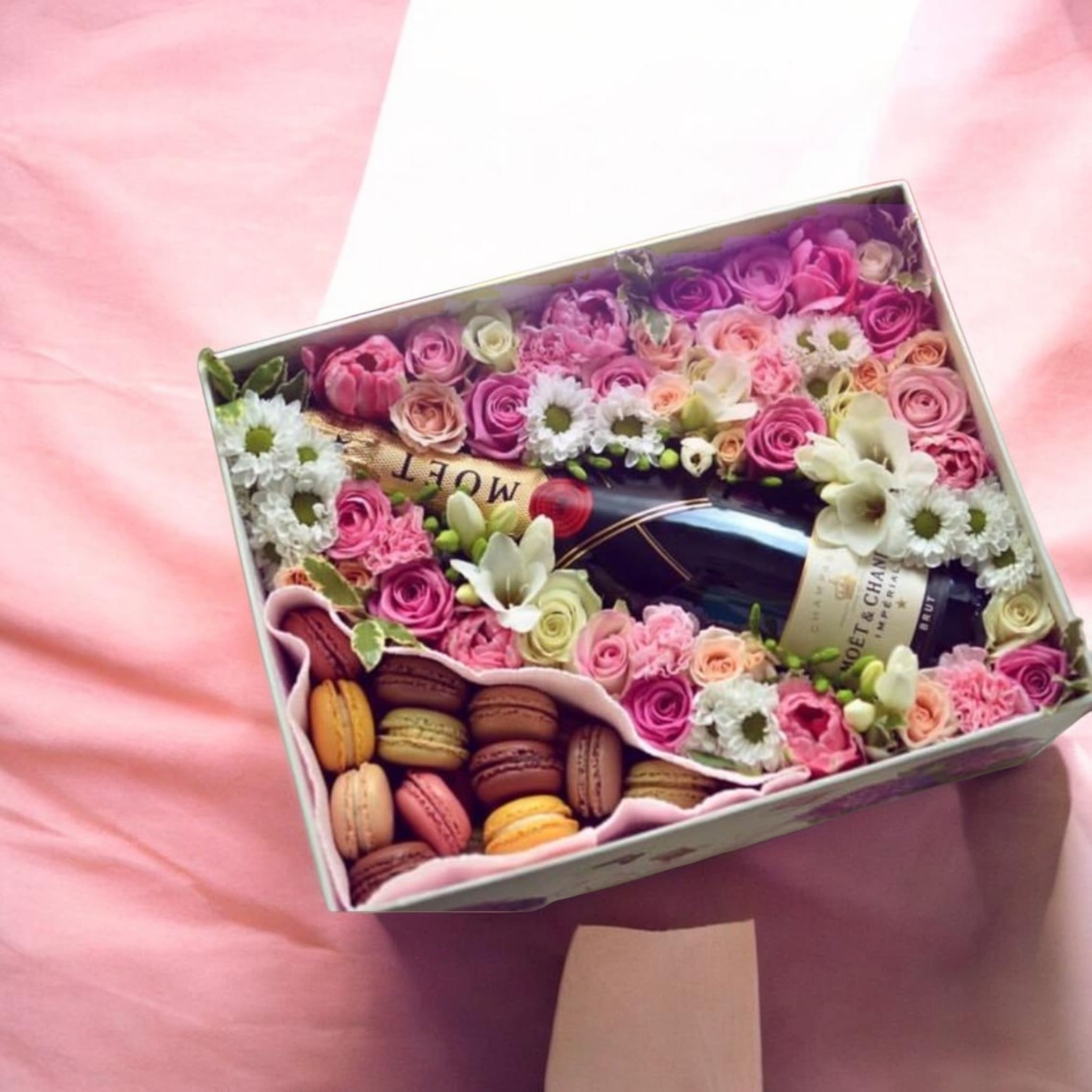 Sofia Pink Rose and Daisy Gift Box Arrangement - A lush cluster of pink roses framed around a chilled bottle of champagne makes this gift-box arrangement a joyful way to mark a birthday, anniversary, or Valentine's Day. The Sofia flower arrangement pairs romantic pink roses with delicate white daisy-like blooms and pale cream accents to create a soft, feminine palette that immediately reads as celebratory and thoughtful for the person you're honoring.  Flowers are densely arranged in a fabric-lined gift box that also holds a tray of assorted macarons, blending floral luxury with a sweet, edible surprise. Hints of purple and yellow among the florals add depth and lift the composition; roses, daisies and complementary filler flowers form a neat decorative frame around the bottle so the presentation looks polished from every angle. The package arrives ready to gift - the champagne is nestled securely, and the macarons sit at the lower corner for an elegant reveal.  Perfect for someone who delights in both blooms and treats, Sofia sends a message of indulgence and care that makes the recipient feel truly celebrated. Choose your size at checkout for proportionate flower counts; keep blooms in a cool spot away from direct sun and enjoy the macarons and champagne within a day or two for peak freshness.