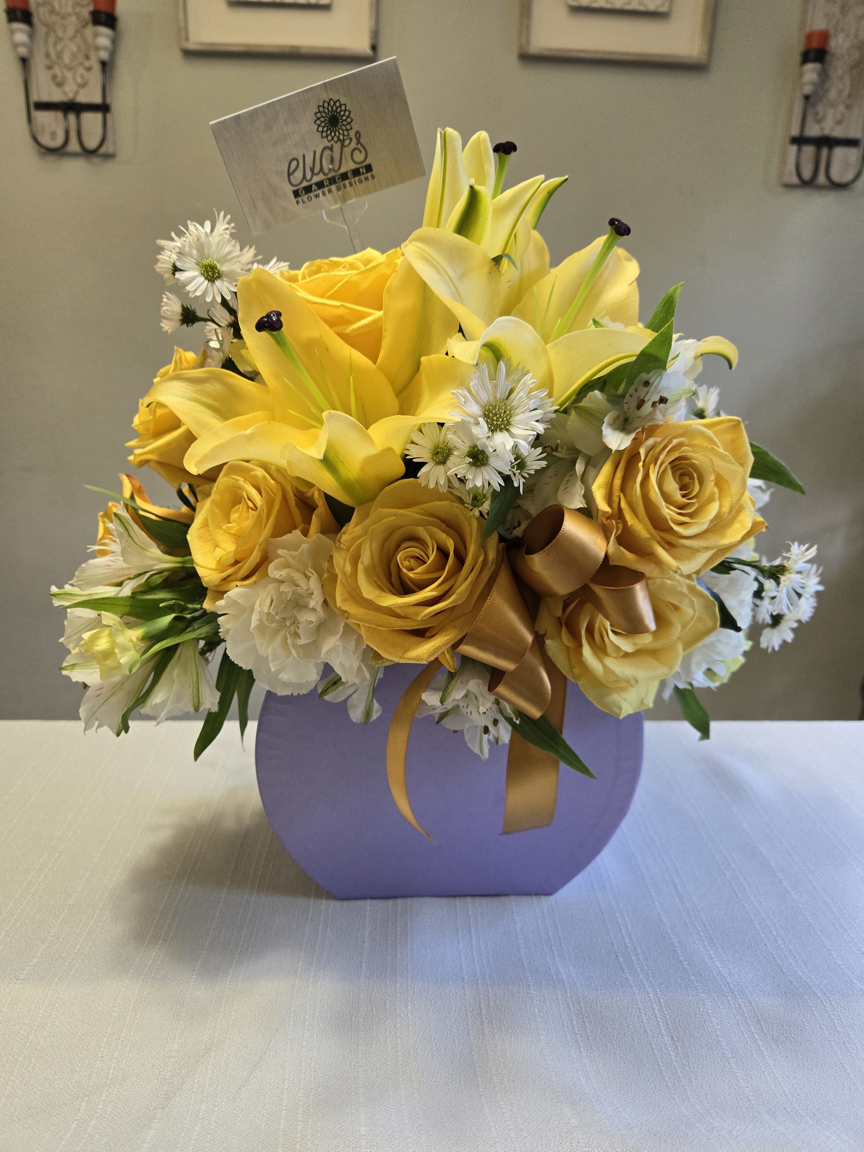 Yellow roses and lilies arranged in a lavender box with a gold ribbon