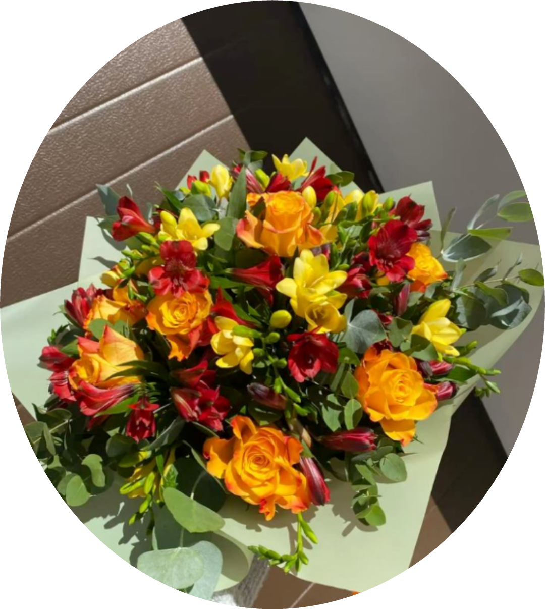 Sunset Drive Orange Rose and Alstroemeria Bouquet - Bright, sunlit color takes center stage in this hand-tied flower arrangement-perfect as a birthday bouquet, a congratulations gift, or any moment you want to make someone feel celebrated. The warm mix of yellow and orange roses paired with red and yellow alstroemeria creates an energetic, cheerful impression that reads as joyful and confident; the recipient will feel noticed and uplifted the moment they see it.  This rounded bouquet features full rose blooms contrasted by the smaller, star-shaped alstroemeria for lively texture and visual interest. Eucalyptus-like foliage threads through the composition, adding soft green texture and a fresh, natural scent. The stems are finished in a pale green bouquet wrap that frames the arrangement and keeps the presentation neat and gift-ready.  Ideal for brightening a mantle, office desk, or kitchen table, this bouquet suits birthdays, congratulations, or simply saying "I'm thinking of you" with flare. Treat it gently: keep stems in fresh water, recut if possible, and place away from direct heat to extend the life of the blooms.