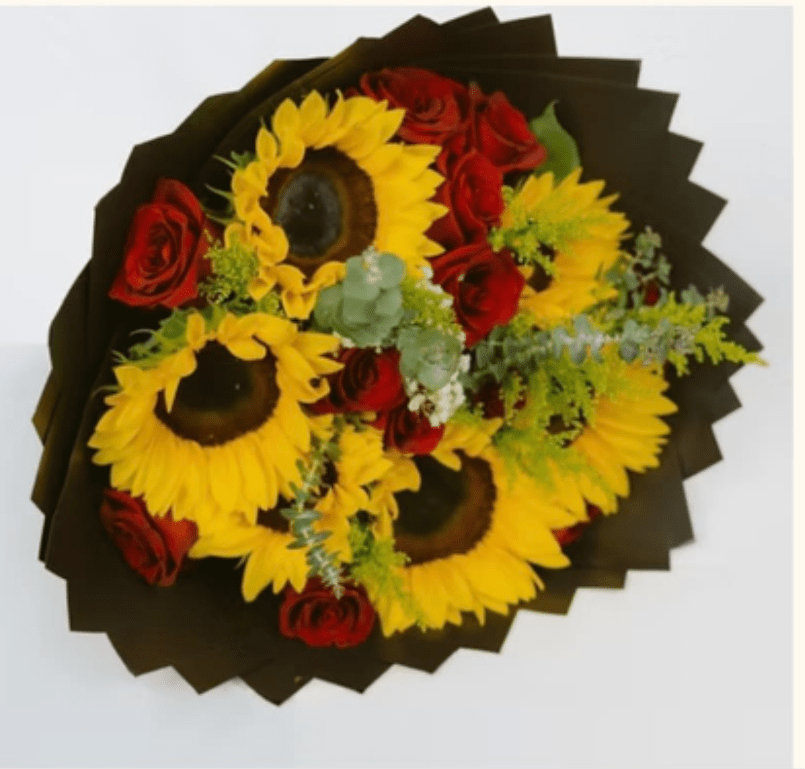 Sunflower and Red Rose Hand-Tied Bouquet - Bright yellow sunflowers paired with clustered red roses create a bold, celebratory birthday bouquet that catches the eye the moment she sees it. The vivid contrast of sunny petals and deep crimson roses against black wrapping paper gives this wrapped bouquet a slightly romantic edge while keeping the mood joyful-perfect for birthdays, congratulations, or any occasion when you want someone to feel seen and celebrated.  This hand-tied arrangement features large sunflowers and multiple red roses arranged in a compact, rounded design with airy green filler to soften edges and add texture. Presented as a wrapped bouquet (no vase), the dark wrapping paper intensifies the colors for a striking visual impact and makes the composition feel modern and confident. Ideal for the person who loves bright, cheerful flowers with a touch of drama-no special care beyond fresh water if transferred to a vase; order today for same-week delivery when available.